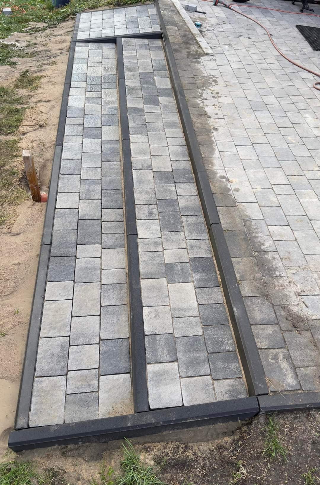 Fragment nowo ułożonej ścieżki z szarej kostki brukowej o różnych odcieniach, obramowanej czarnym krawężnikiem. Widoczny piasek po bokach i fragment istniejącego bruku w tle.