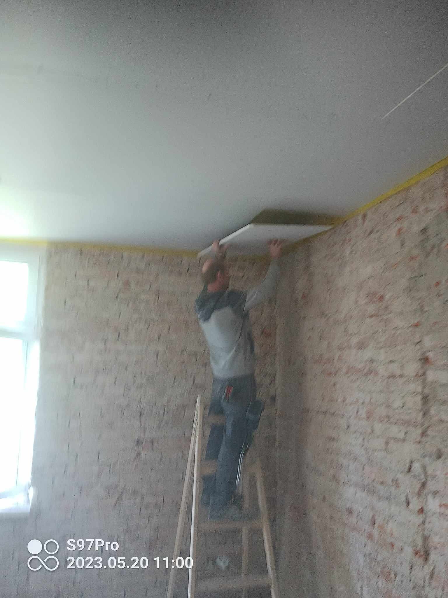 Pracownik na drabinie montuje płytę kartonowo-gipsową na suficie w pomieszczeniu z surowymi ścianami z cegły, zabezpieczonymi żółtą taśmą malarską.