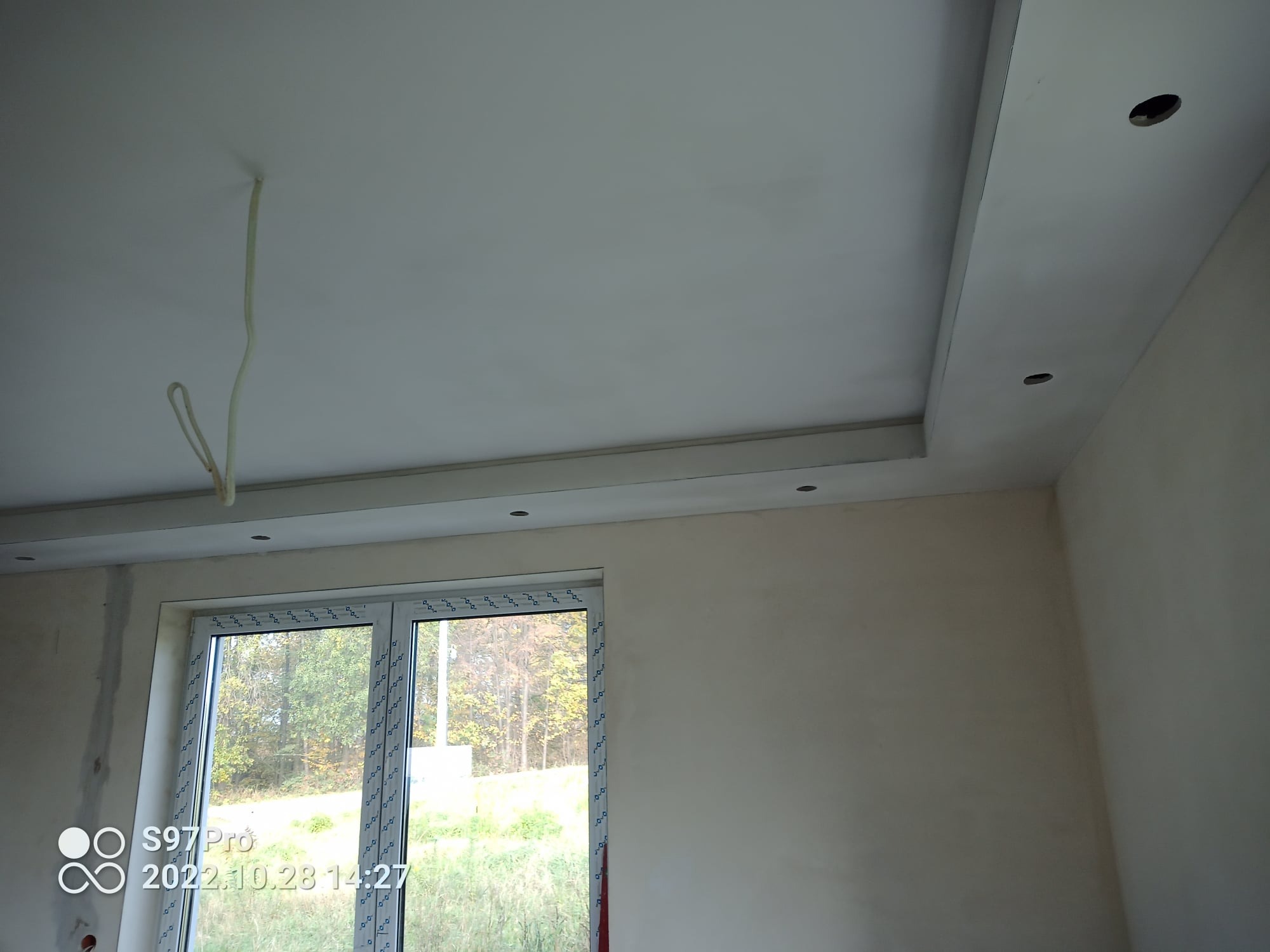 Wykończony sufit podwieszany z oświetleniem punktowym w nowym pomieszczeniu, widoczne wystające przewody elektryczne i fragment okna z widokiem na drzewa.