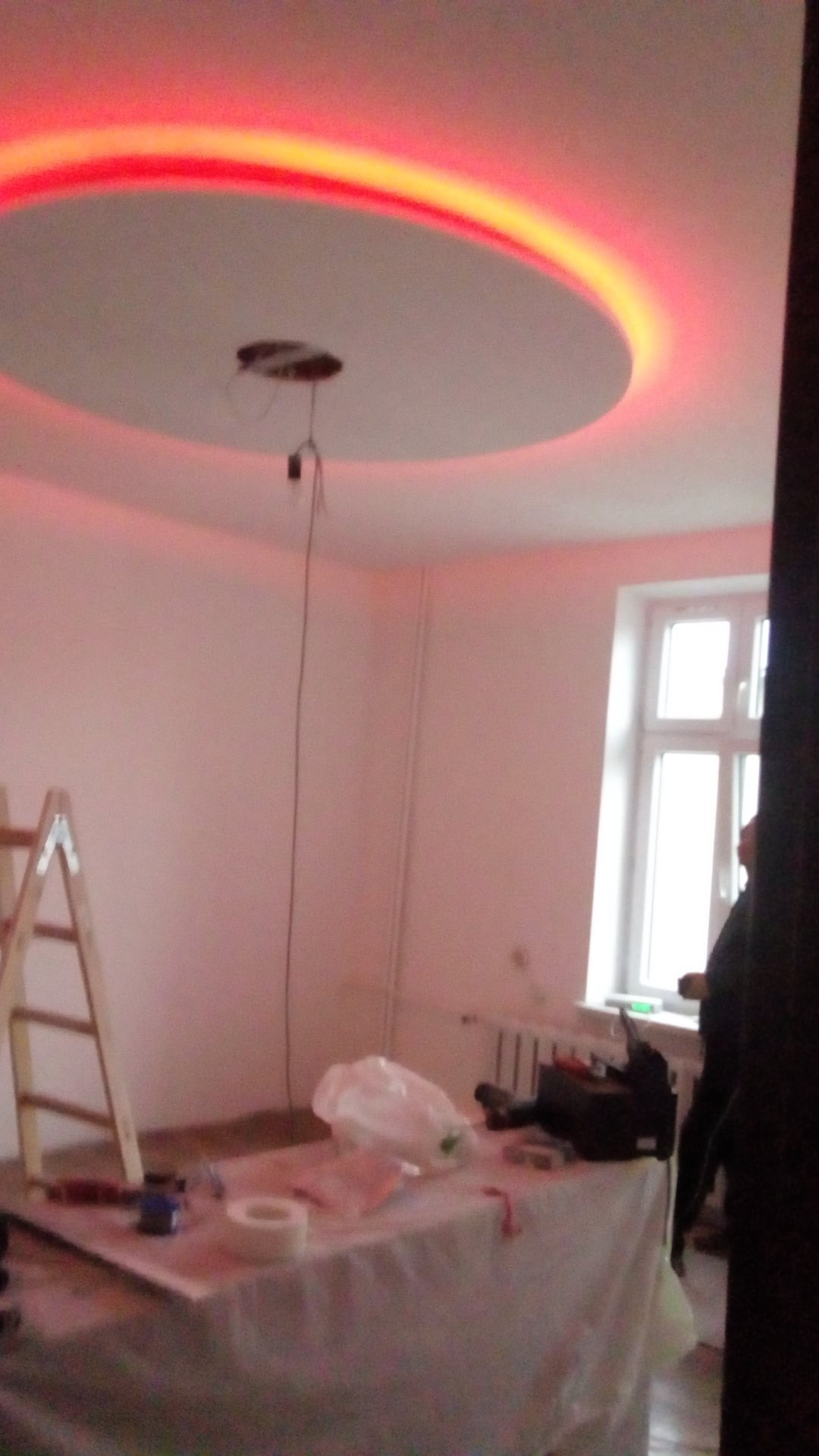 Niedokończony sufit podwieszany z okrągłym wgłębieniem oświetlonym czerwoną listwą LED, drabina oparta o ścianę, stolik z narzędziami i materiałami budowlanymi w pokoju z oknem.