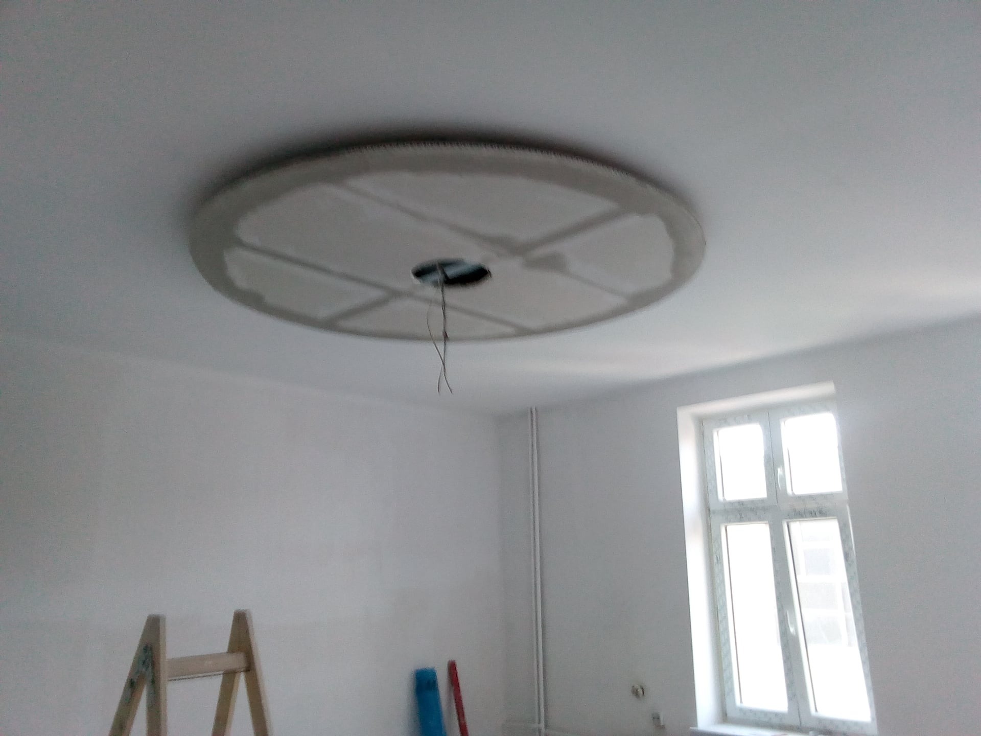 Okrągła konstrukcja z płyty kartonowo-gipsowej na suficie, z otworem na oświetlenie i widocznymi przewodami, w trakcie prac wykończeniowych w pomieszczeniu z drabiną i oknem.