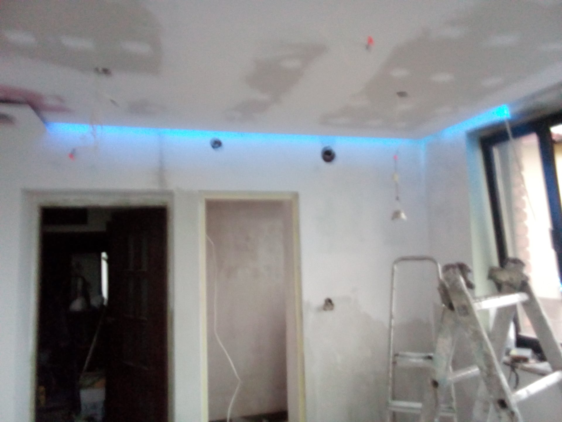 Wnętrze w trakcie prac przygotowawczych: ściany po szpachlowaniu, sufit z widocznymi otworami na oświetlenie i zainstalowanym niebieskim oświetleniem LED, aluminiowa drabina w rogu pokoju.