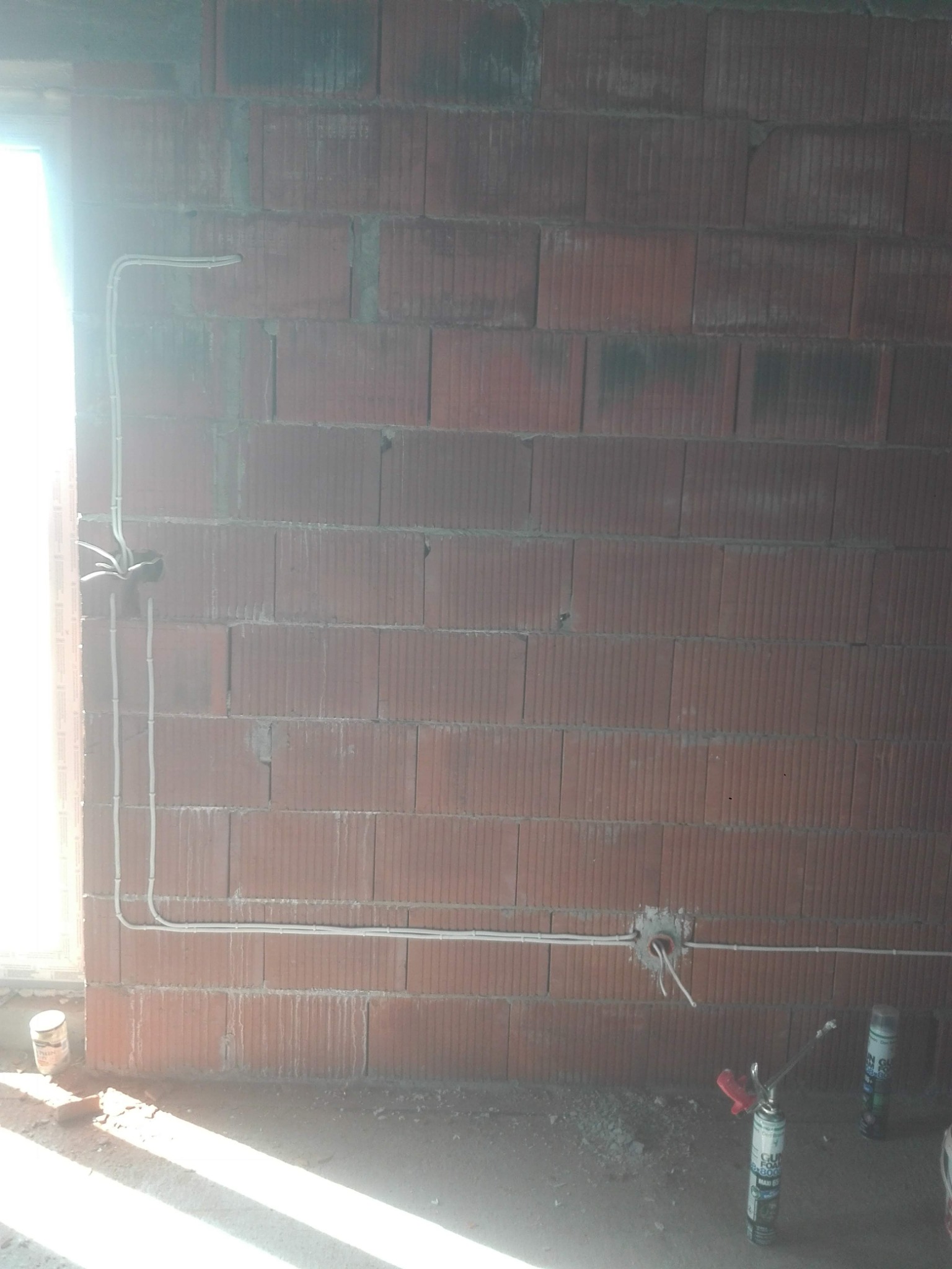 Ściana z czerwonej cegły z ułożonymi przewodami elektrycznymi, puszka instalacyjna z wystającymi kablami, pianka montażowa w tle.