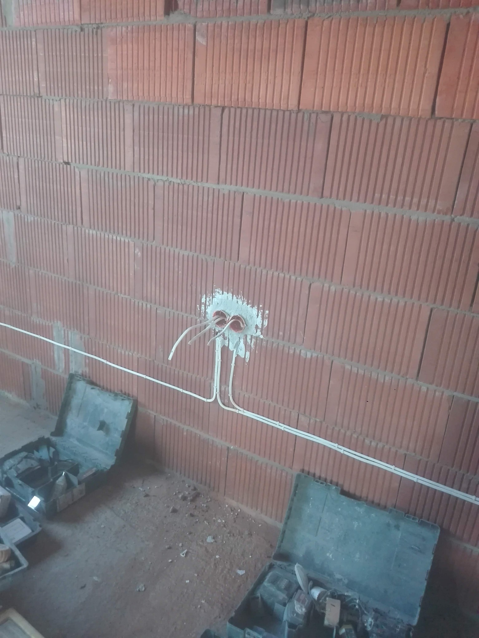 Czerwona ściana z pustaków ceramicznych z widocznymi przewodami elektrycznymi wychodzącymi z puszki instalacyjnej; na podłodze rozłożone skrzynki narzędziowe.