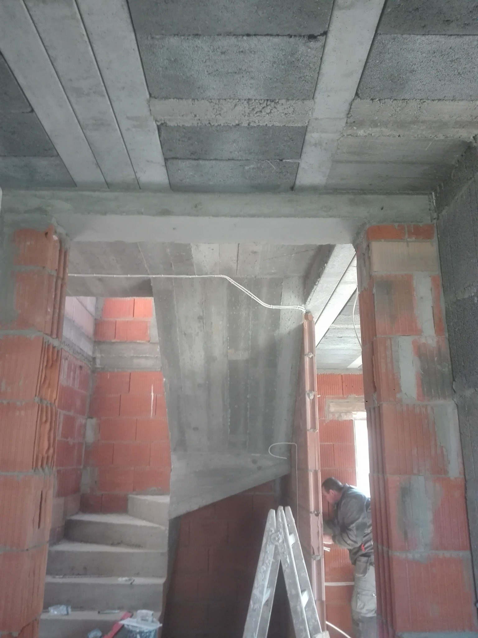 Wnętrze w trakcie prac budowlanych: surowy betonowy strop z widocznymi belkami, ściany z czerwonej cegły, schody w szarym betonie oraz pracownik na drabinie instalujący przewody elektryczne.
