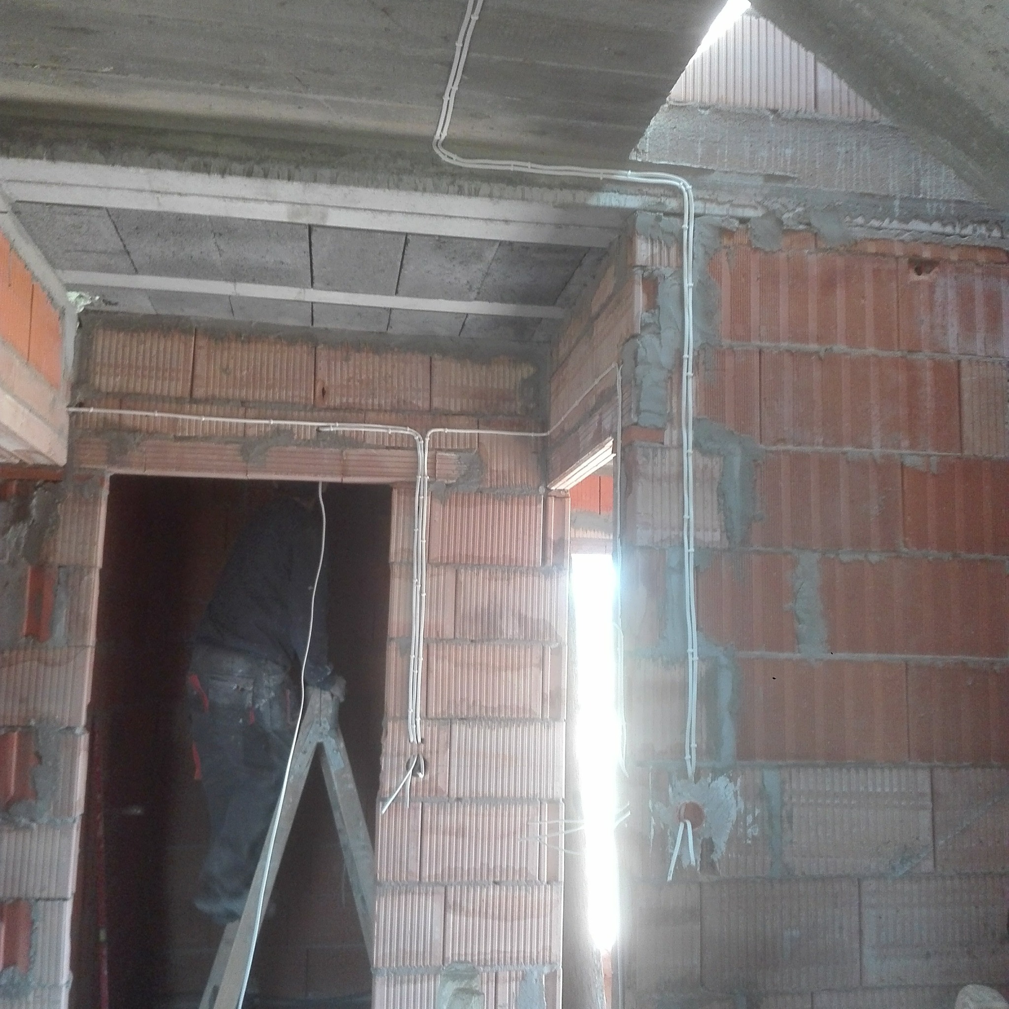 Instalacja elektryczna w surowym stanie: białe przewody prowadzone po ceglanej ścianie, częściowo zamurowane, widoczny pracownik na drabinie w tle.