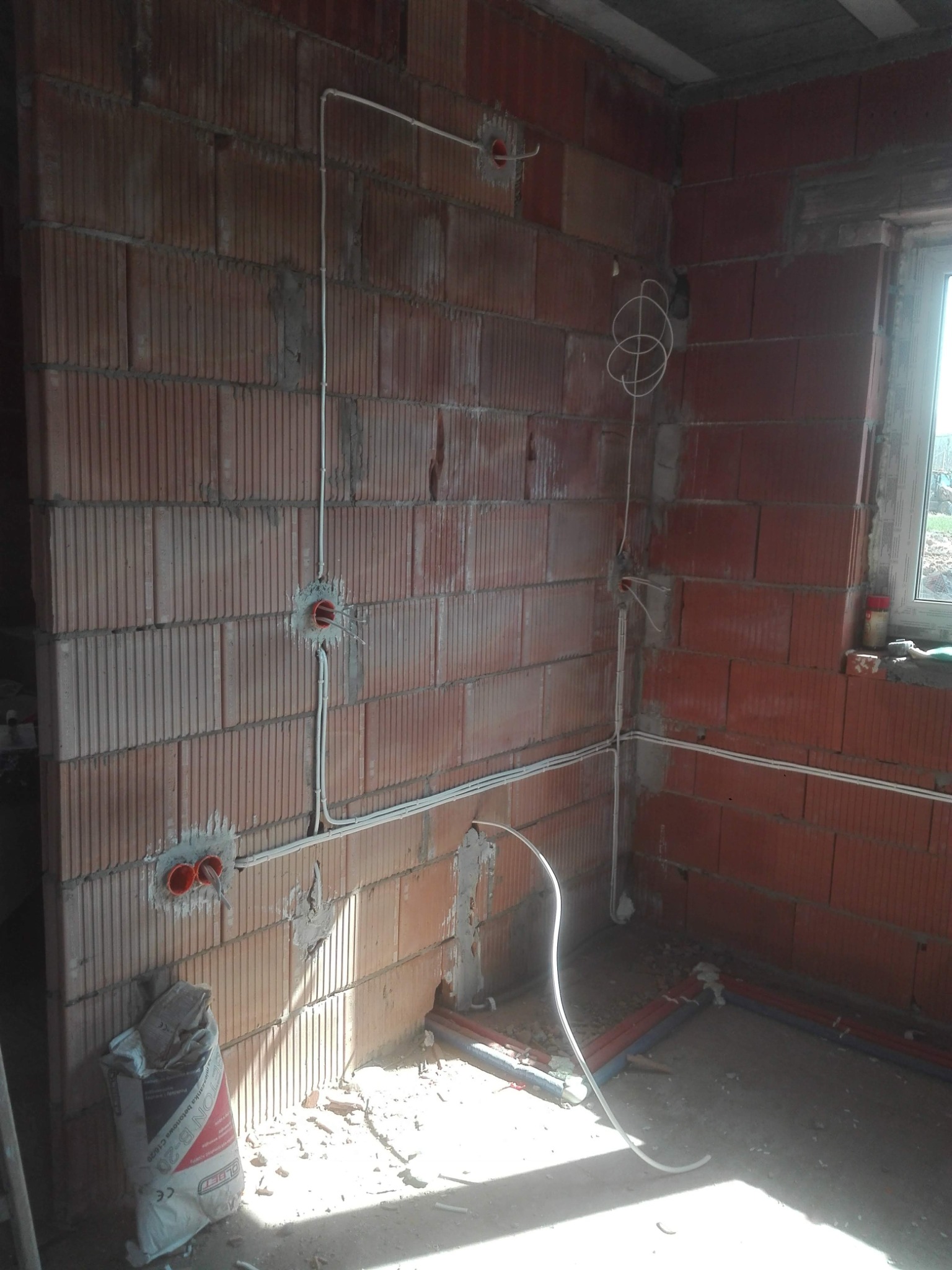 Instalacja elektryczna w surowym pomieszczeniu z cegły: białe kable poprowadzone po ścianach, puszki elektryczne osadzone w murze, worek z betonem przy podłodze.