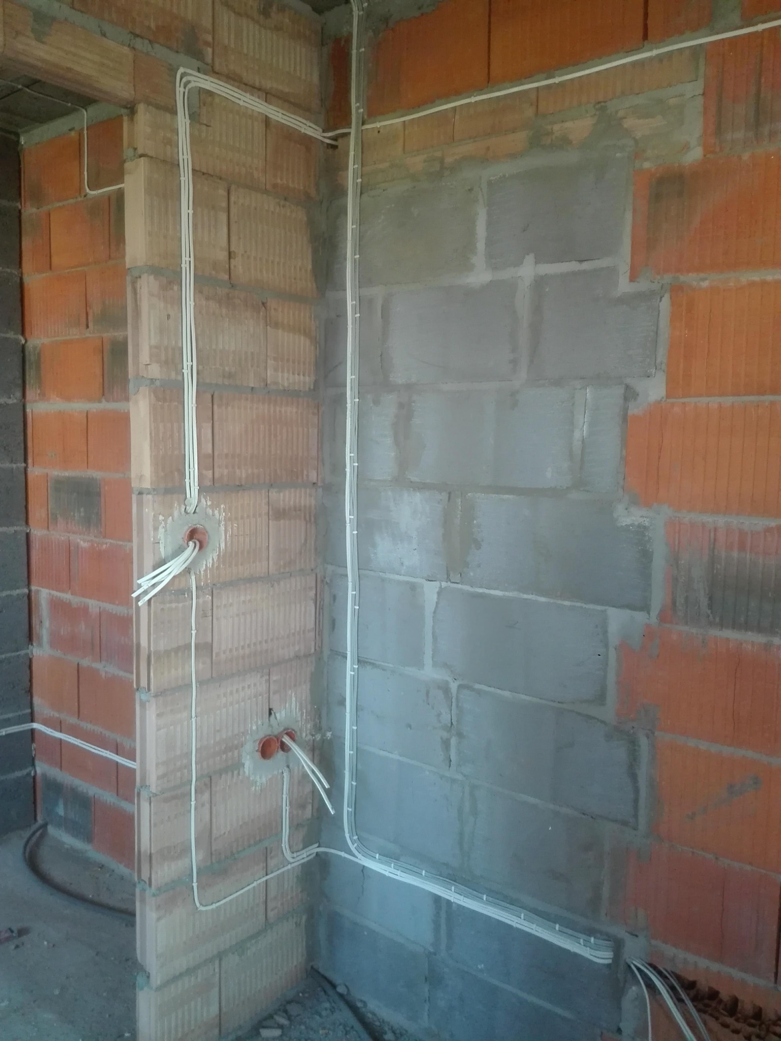 Surowe ściany z cegły i betonu z poprowadzonymi natynkowymi przewodami elektrycznymi w białych listwach, widoczne puszki instalacyjne z wystającymi kablami.