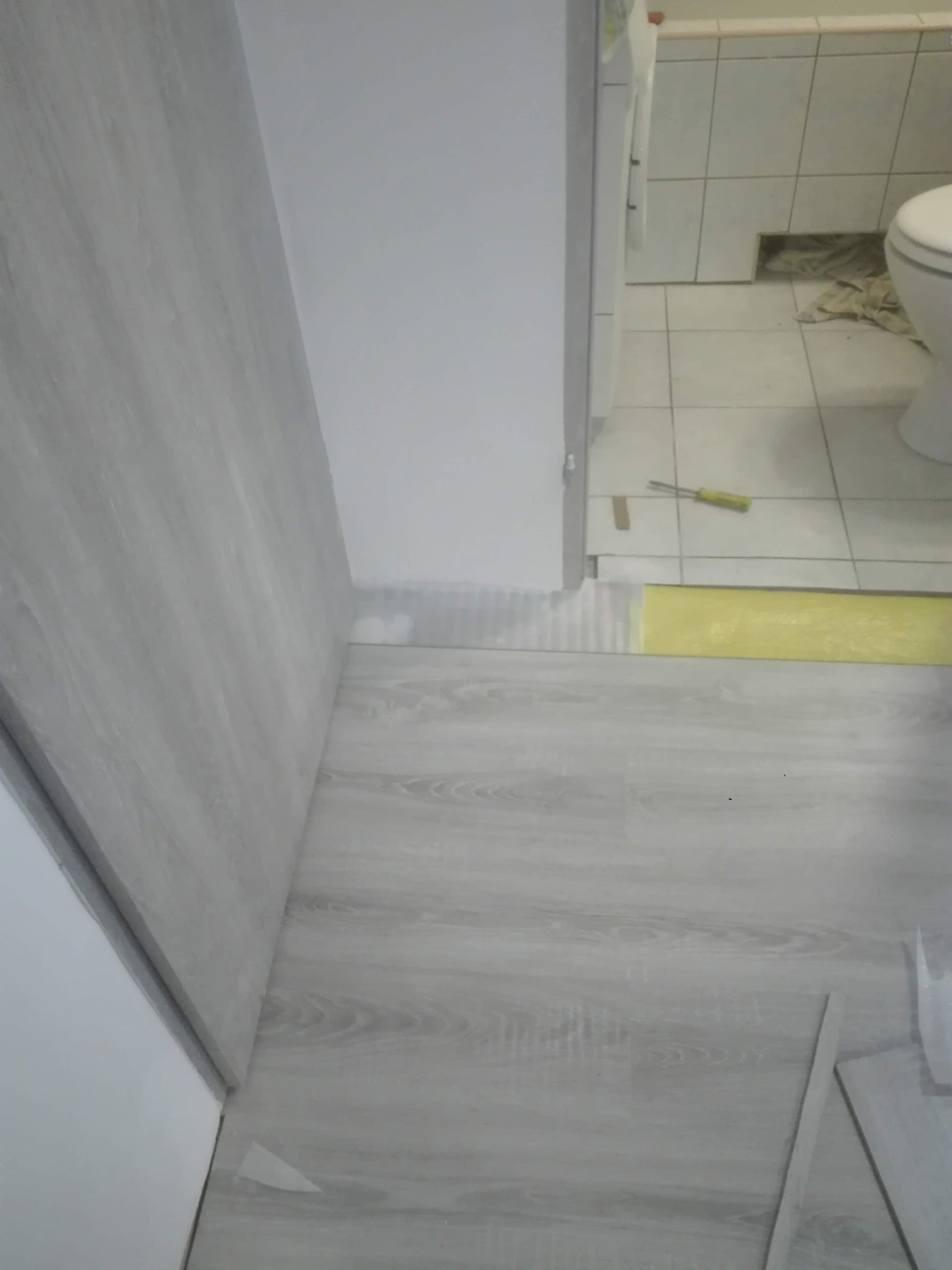 Układanie paneli podłogowych w łazience, widoczny fragment toalety i płytki na ścianie, żółta taśma zabezpieczająca.