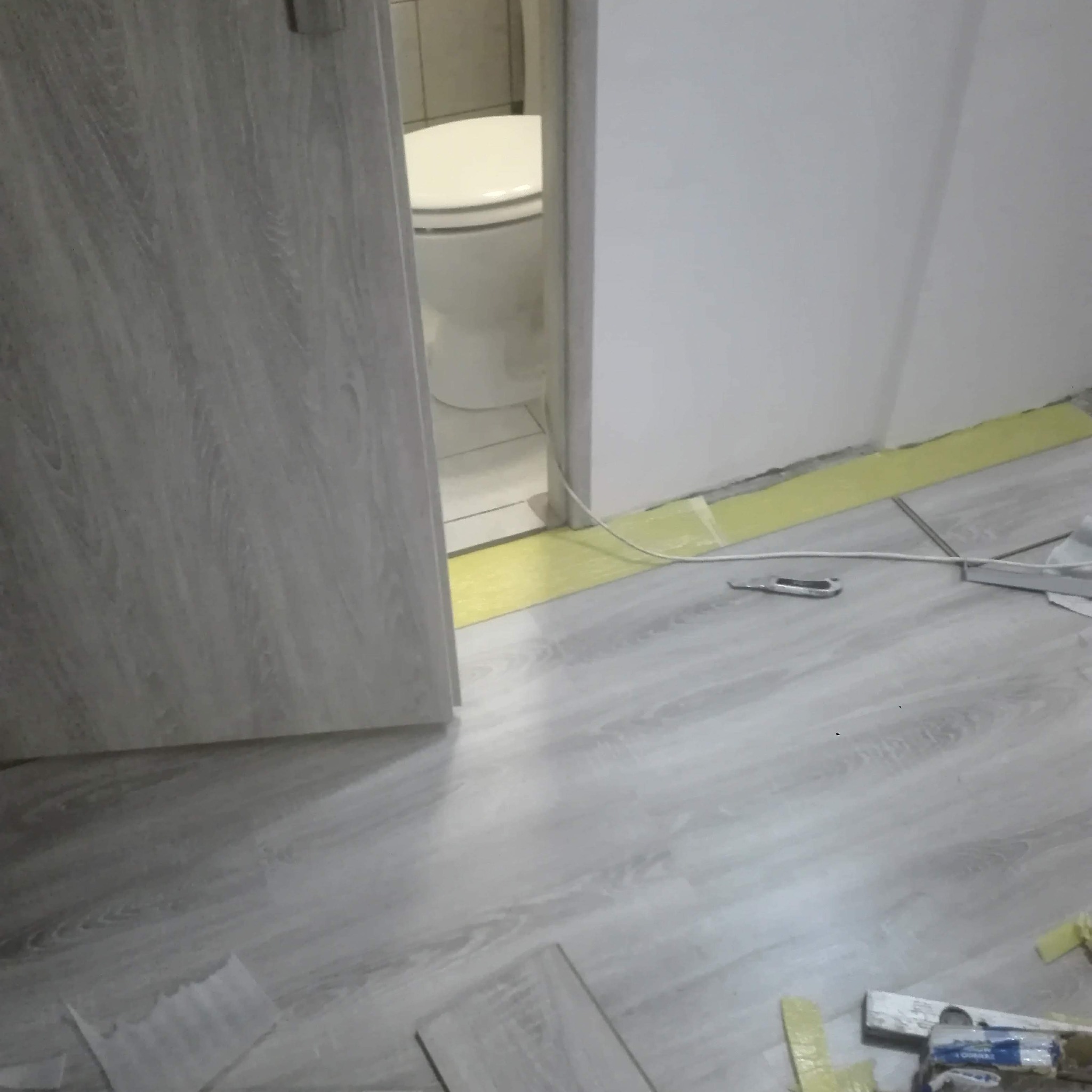 Układanie paneli podłogowych w łazience z widoczną toaletą, narzędzia pracy i żółta taśma maskująca przy ścianie.