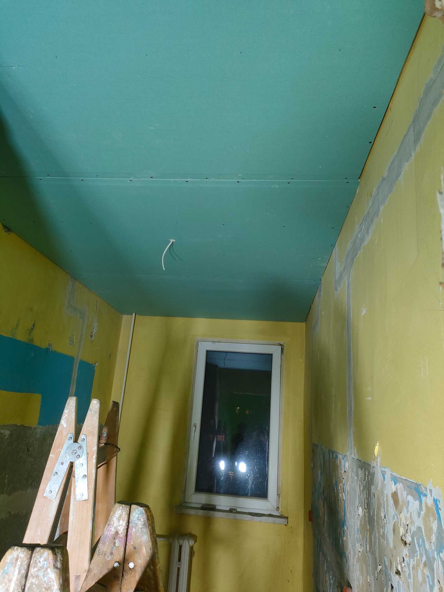 Montaż zielonej płyty karton-gips na suficie w pokoju z żółtymi ścianami, widoczna drabina malarska i fragment instalacji elektrycznej.