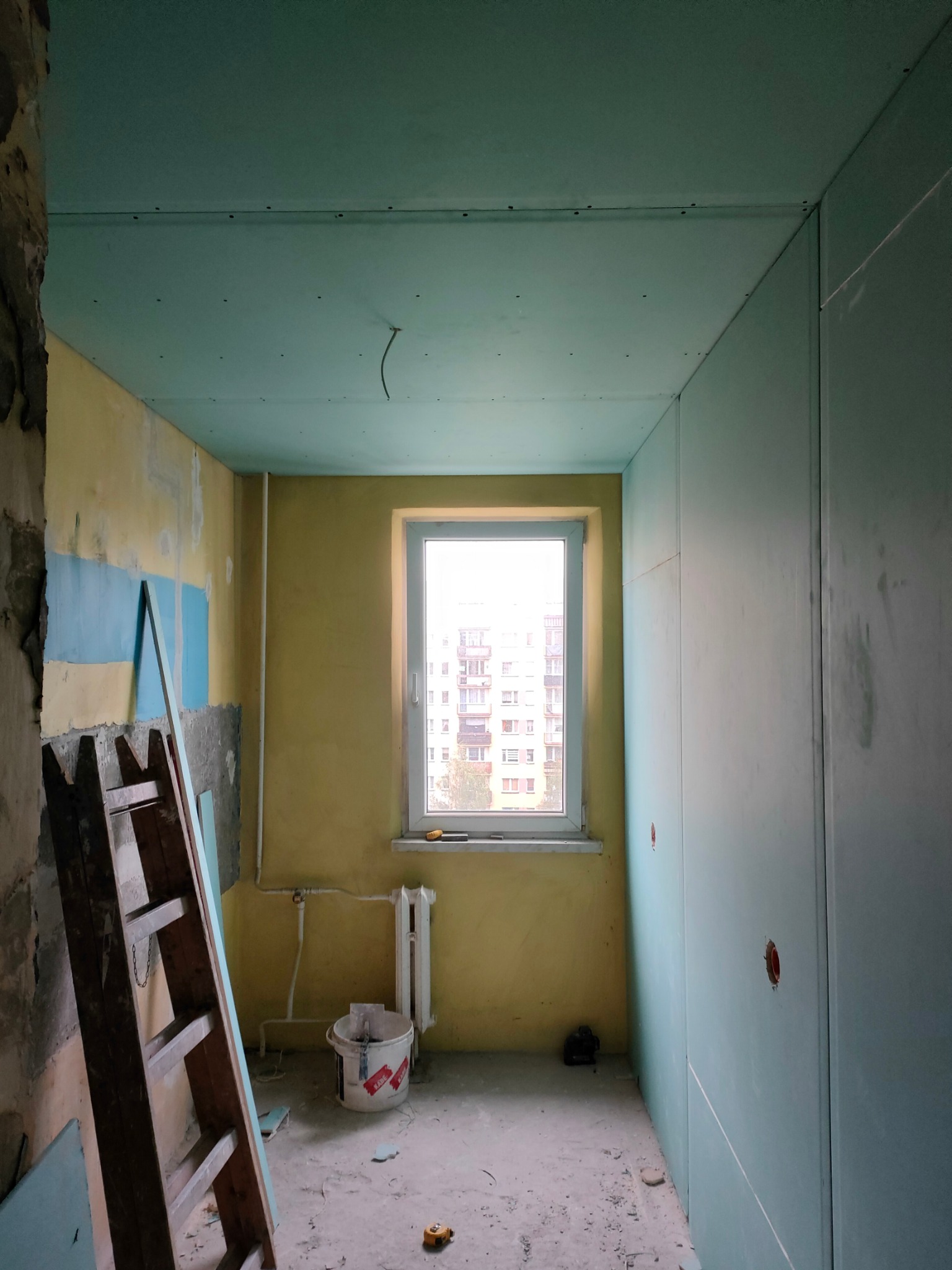 Pokój w trakcie remontu: ściany i sufit obłożone niebieskimi płytami kartonowo-gipsowymi, widoczna drewniana drabina oparta o ścianę z fragmentami starej farby, okno z widokiem na blok, na podłodze...