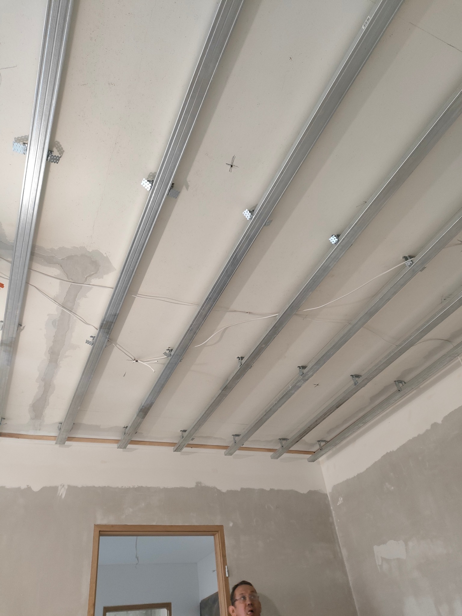 Metalowa konstrukcja sufitu podwieszanego z płyt kartonowo-gipsowych, widoczne profile i uchwyty montażowe, fragment instalacji elektrycznej w tle, surowa ściana przygotowana do dalszych prac...