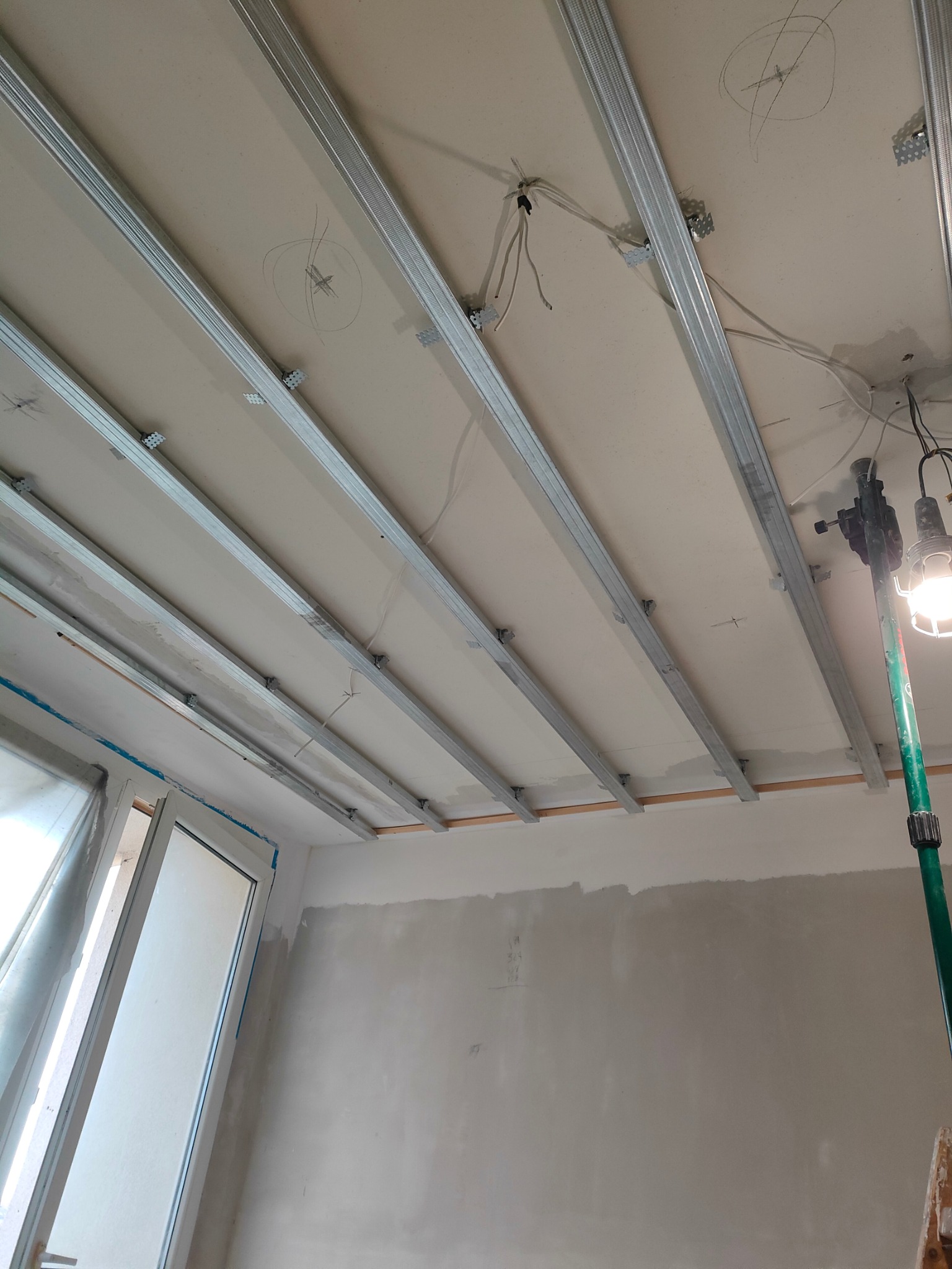 Stelaż sufitu podwieszanego z płyt kartonowo-gipsowych, widoczne profile metalowe, instalacja elektryczna w suficie oraz lampa robocza na statywie.