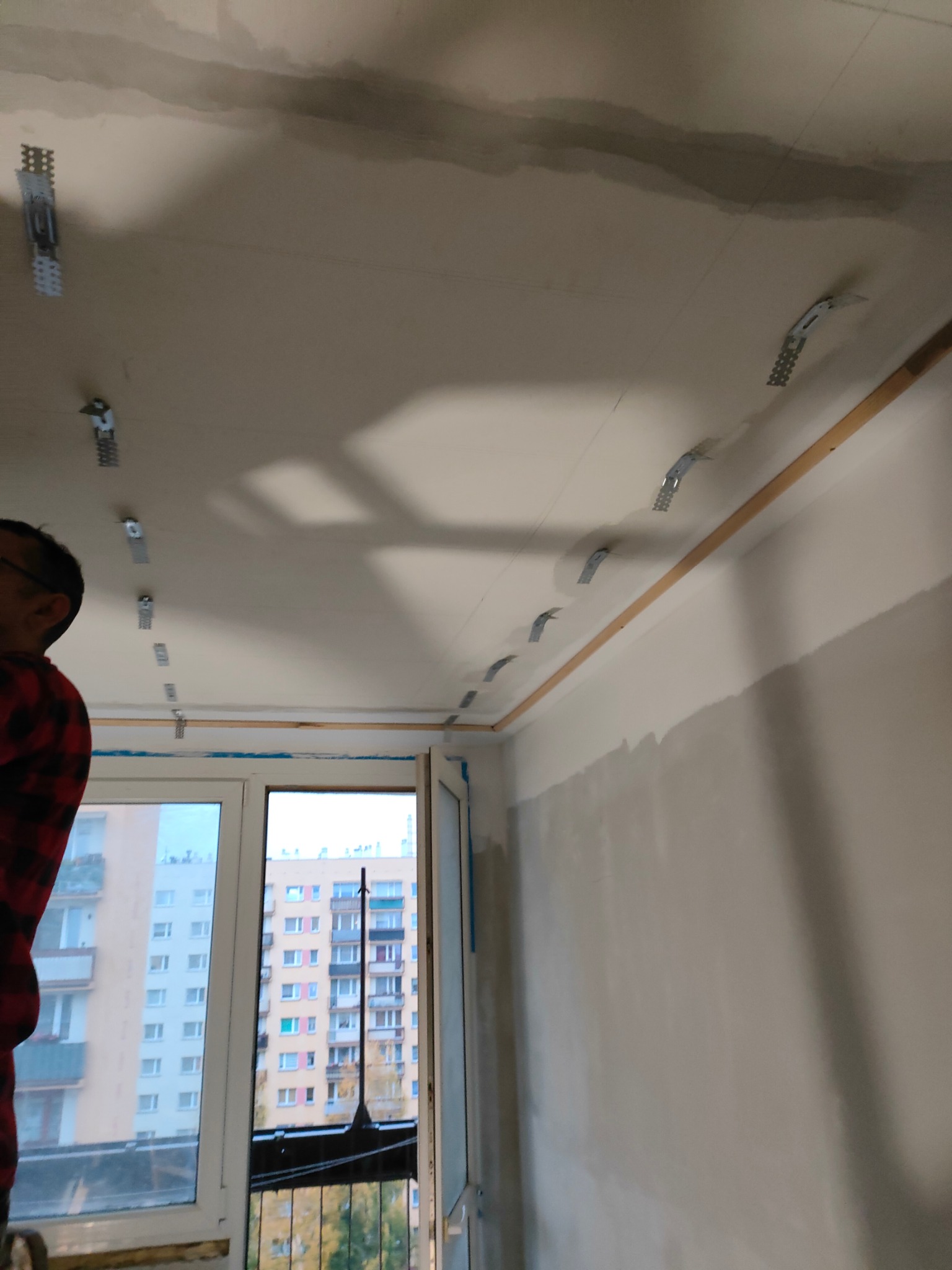 Montaż metalowych profili do sufitu podwieszanego, widoczne profile mocujące, listwa przypodłogowa, okno z widokiem na blok mieszkalny w tle, fragment sylwetki osoby w czerwonej koszuli w kratę.