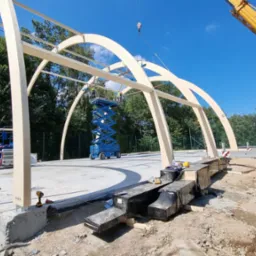 Montaż drewnianej konstrukcji łukowej hali sportowej z użyciem dźwigu i podnośnika nożycowego na betonowej płycie fundamentowej.
