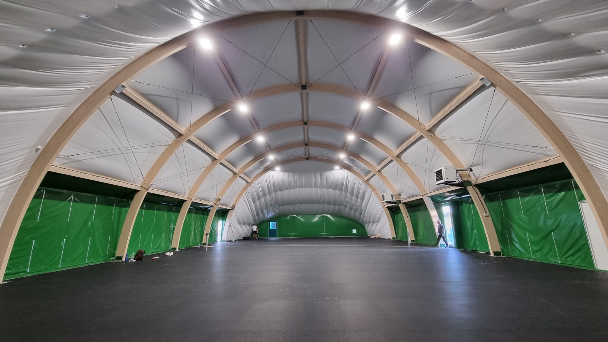 Wnętrze hali sportowej z drewnianą konstrukcją łukową, oświetleniem LED i zielonymi ścianami bocznymi. Widoczna osoba w oddali.