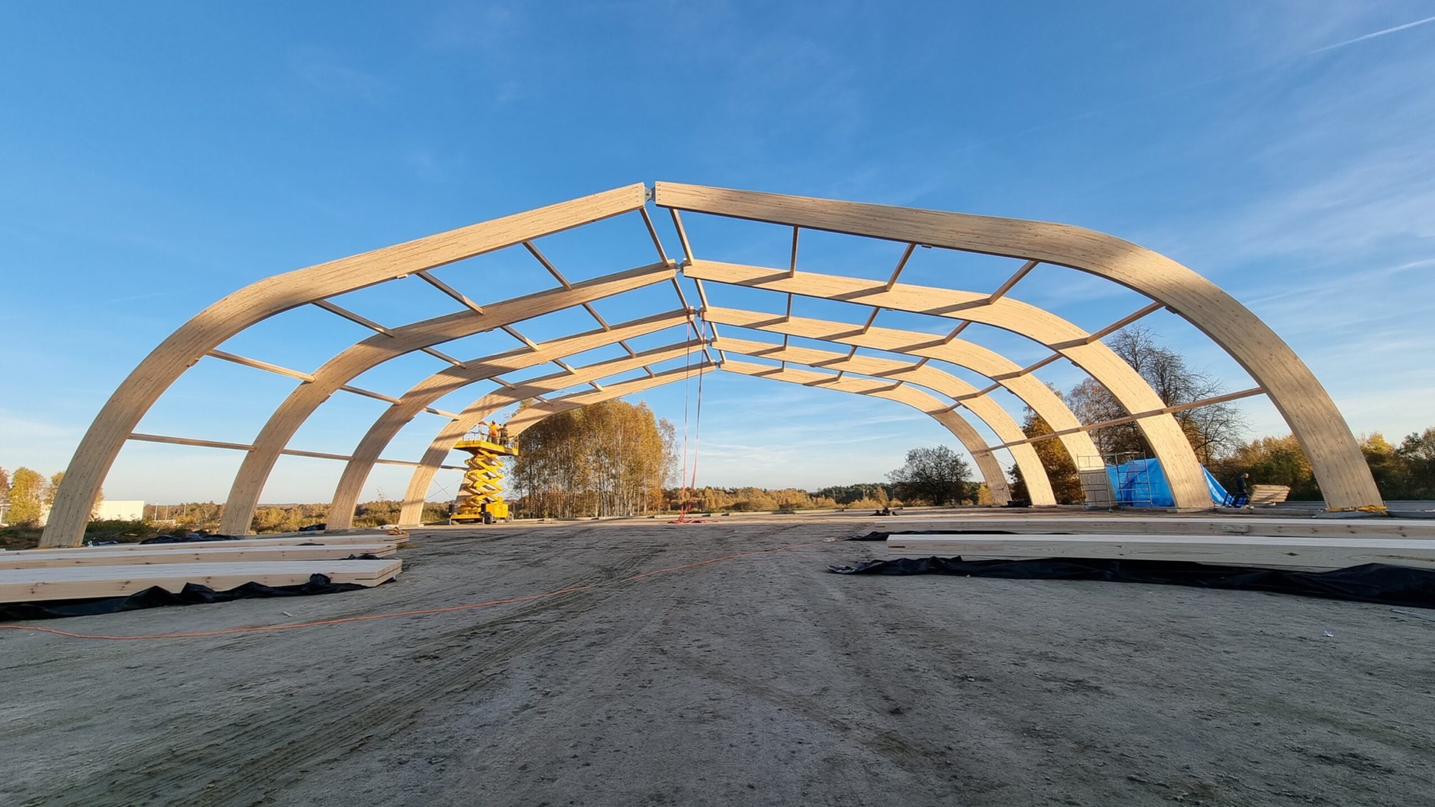 Wznoszenie drewnianej konstrukcji łukowej hali na placu budowy, widoczny podnośnik koszowy i przygotowane elementy drewniane.