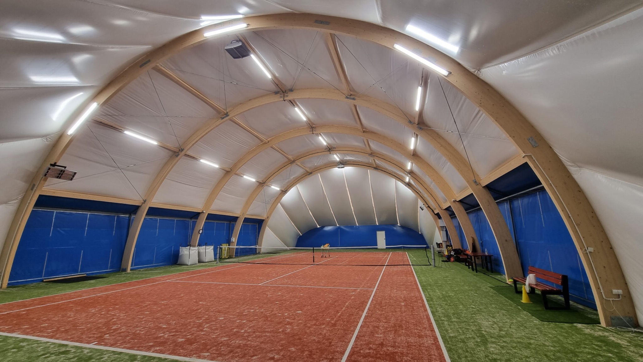 Wnętrze hali sportowej z drewnianą konstrukcją łukową, oświetleniem LED i kortem tenisowym z czerwoną nawierzchnią.