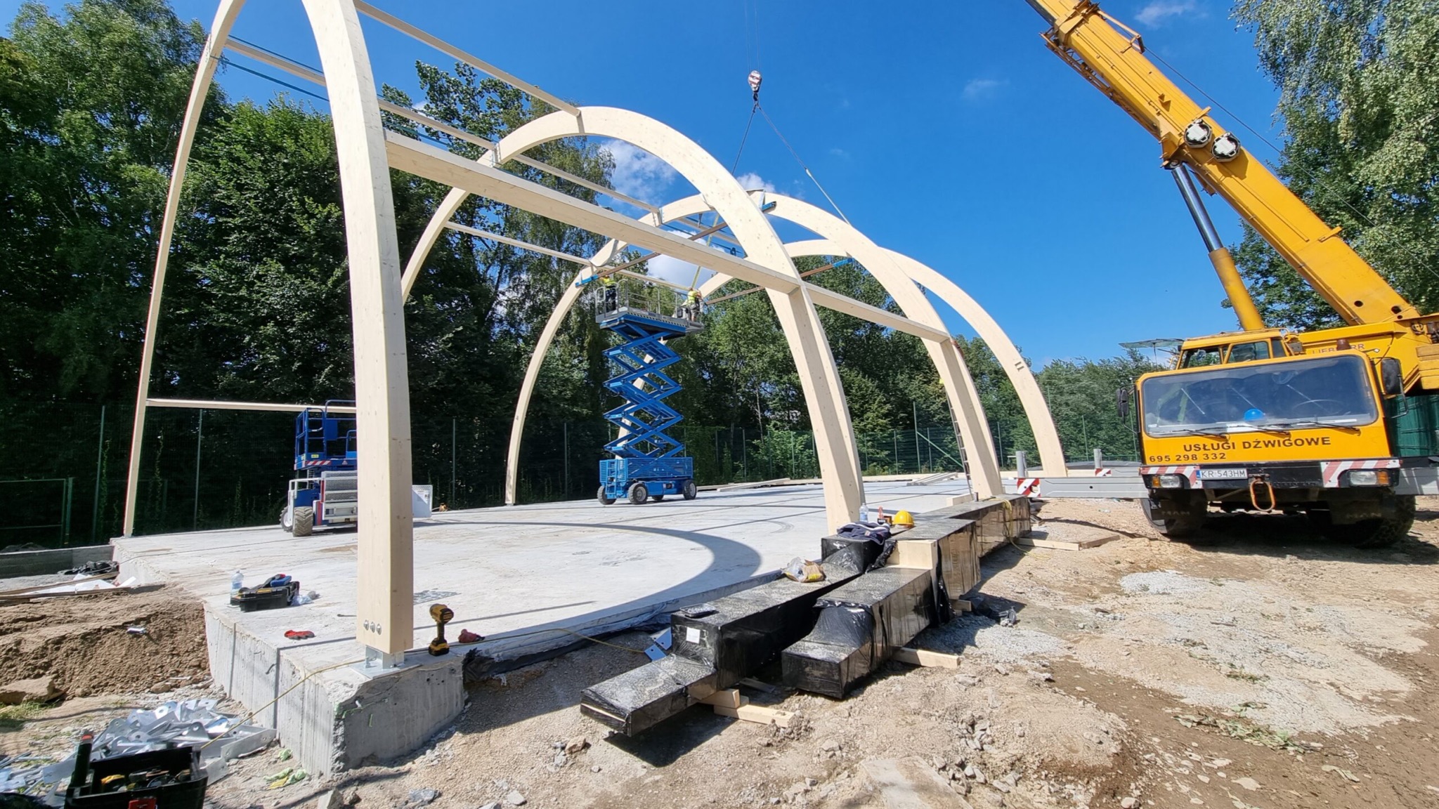 Montaż drewnianej konstrukcji łukowej hali sportowej z użyciem dźwigu i podnośnika nożycowego na betonowej płycie fundamentowej.