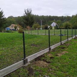 ARTPRO OGRODZENIA - Montaż ogrodzenia panelowego z podmurówką betonową na trawiastym terenie wiejskim, widoczne ślady prac ziemnych.