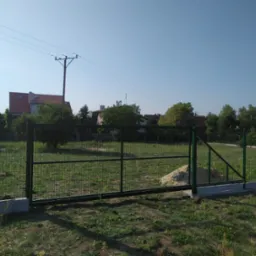Nowo zainstalowana, zielona brama wjazdowa z metalowej siatki na betonowej podmurówce, widok z zewnątrz posesji, w tle domy i słup elektryczny.