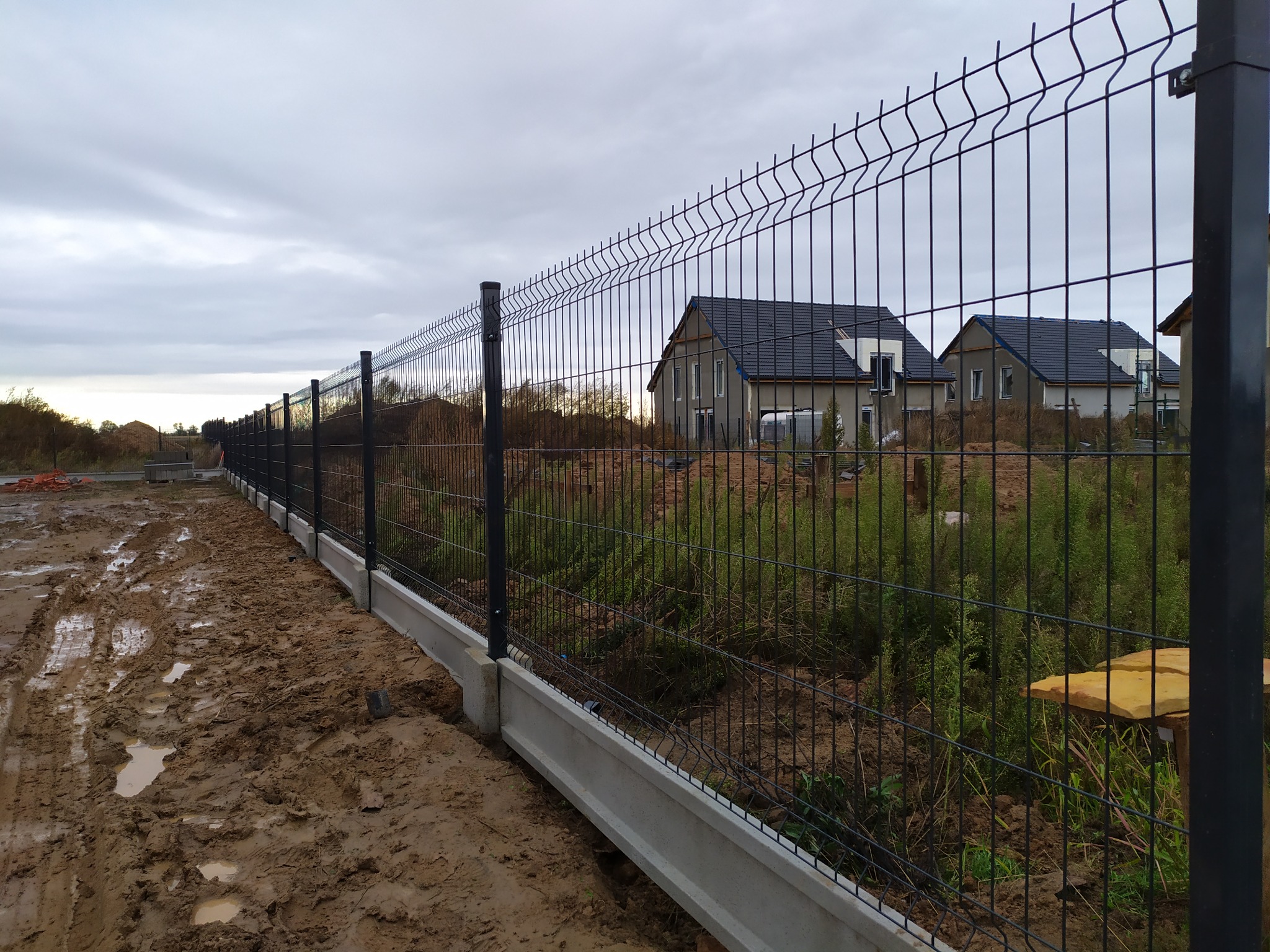 Długi, czarny panelowy płot z betonową podmurówką wzdłuż błotnistej ścieżki, z domami w tle pod pochmurnym niebem.