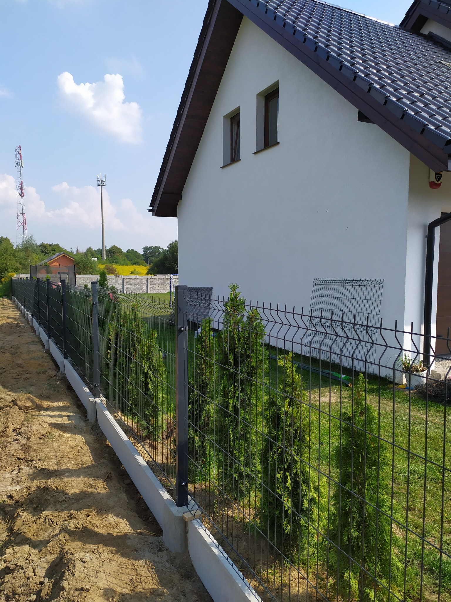 Nowoczesne ogrodzenie panelowe z podmurówką betonową, obsadzone tujami, na tle domu z brązową dachówką i zielenią