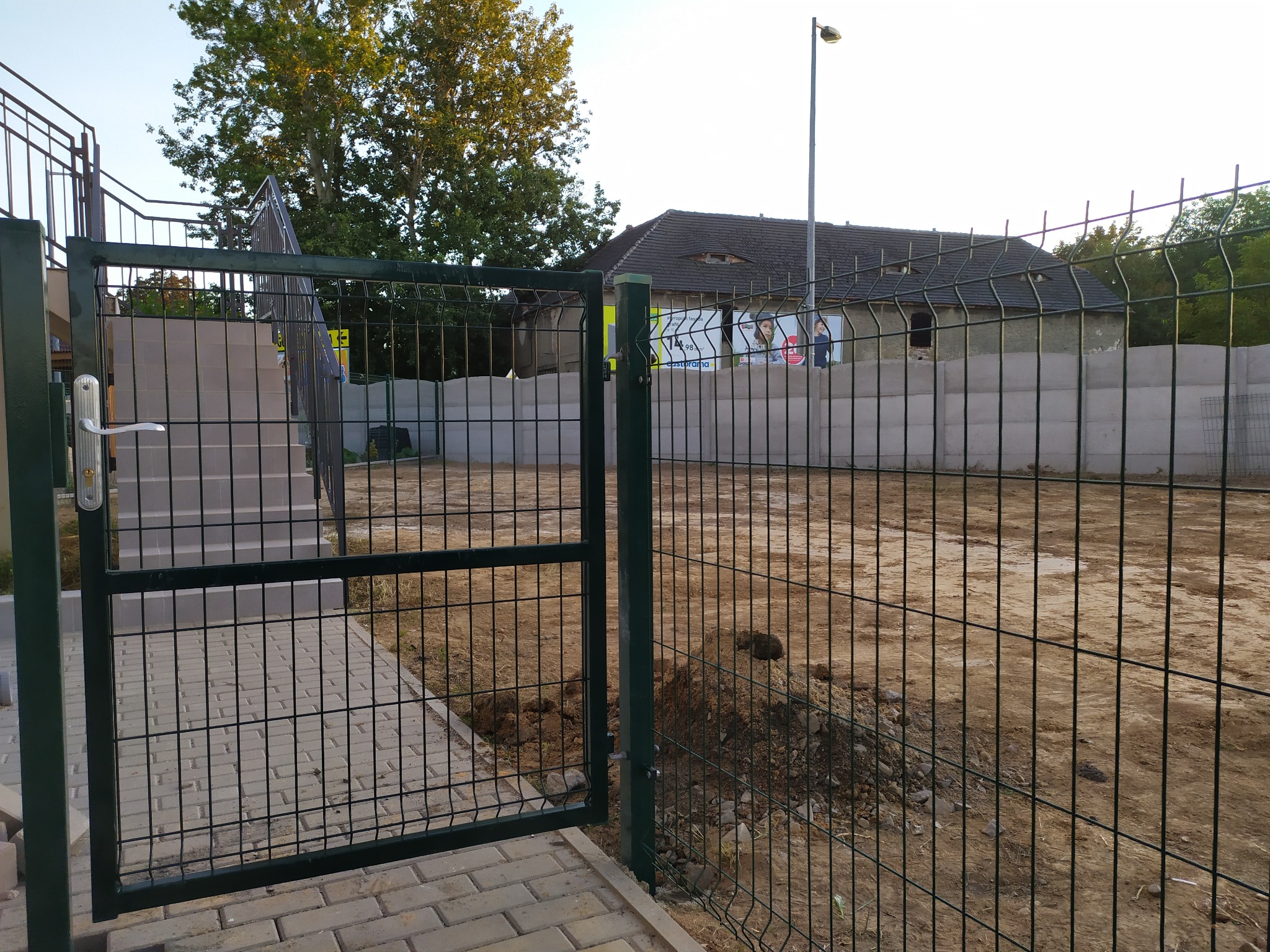 Otwarta furtka z metalowej siatki ogrodzeniowej na posesję z nierównym terenem, w tle budynek z betonowych bloków i reklamy.