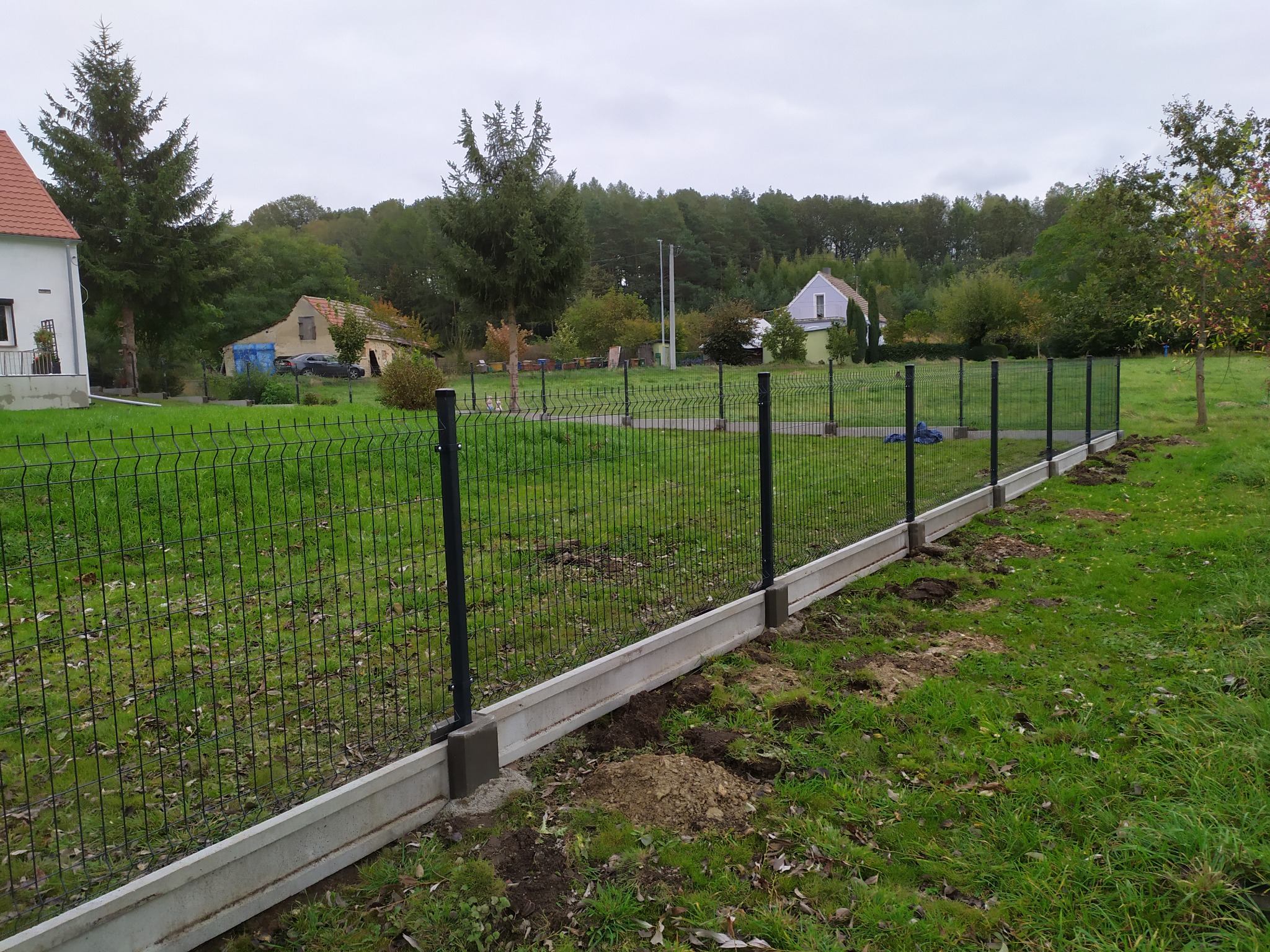 Montaż ogrodzenia panelowego z podmurówką betonową na trawiastym terenie wiejskim, widoczne ślady prac ziemnych.