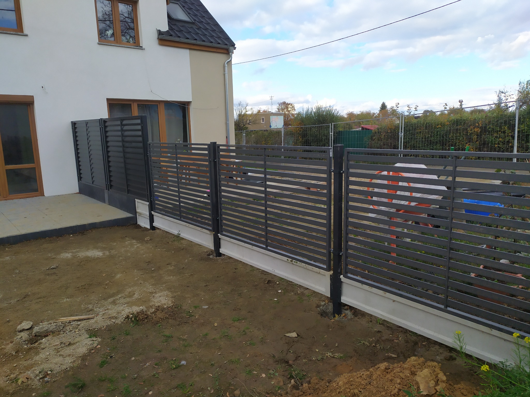 Nowoczesne, poziome, szare ogrodzenie z metalowych paneli, zamontowane na betonowej podmurówce, widoczne na tle jasnego domu i częściowo przygotowanego terenu.