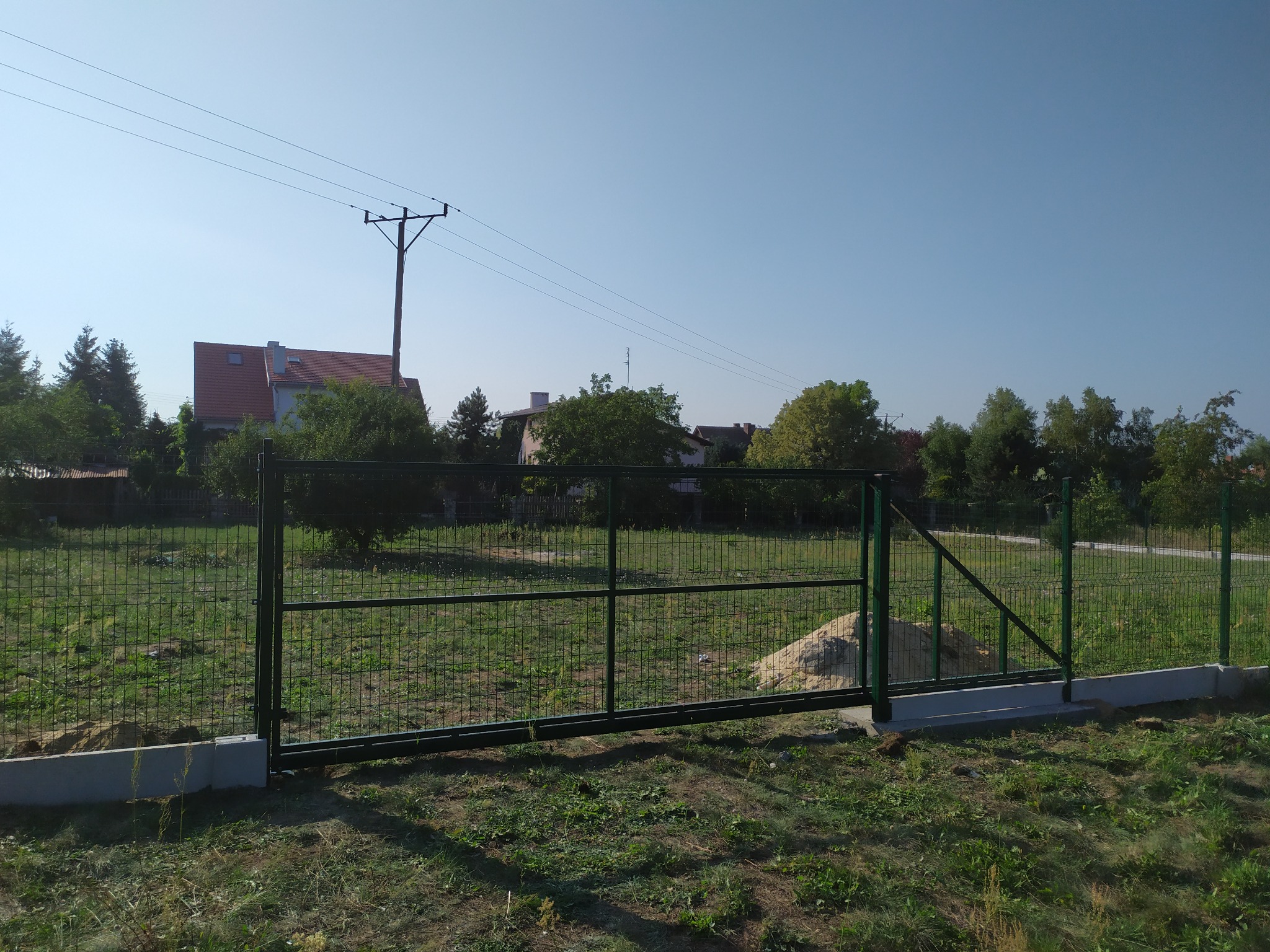 Nowo zainstalowana, zielona brama wjazdowa z metalowej siatki na betonowej podmurówce, widok z zewnątrz posesji, w tle domy i słup elektryczny.