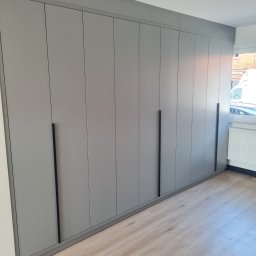 MEBLE-SKIBA TOMASZ SKIBA - Szara zabudowa meblowa od podłogi do sufitu z minimalistycznymi, pionowymi uchwytami w czarnym kolorze, na tle jasnej podłogi imitującej drewno i okna z widokiem na ulicę.