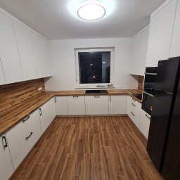 MEBLE-SKIBA TOMASZ SKIBA - Nowoczesna kuchnia w kształcie litery U z białymi szafkami, drewnianym blatem i podłogą imitującą drewno, oświetlona centralnym plafonem.