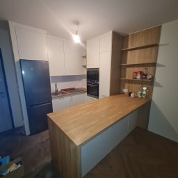 MEBLE-SKIBA TOMASZ SKIBA - Nowoczesna kuchnia z białymi szafkami, drewnianym blatem i wyspą kuchenną, widok z perspektywy podłogi, z otwartymi półkami na ścianie. Na blacie stoją przyprawy i napoje.