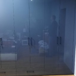 MEBLE-SKIBA TOMASZ SKIBA - Szafa wnękowa z ciemnymi, lakierowanymi frontami, widoczne odbicie sylwetki osoby, szuflady w dolnej części, parkiet na podłodze.