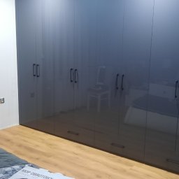 MEBLE-SKIBA TOMASZ SKIBA - Duża, ciemnoszara szafa wnękowa z połyskującymi frontami i podłużnymi uchwytami, widoczna w sypialni z łóżkiem zagraconym pudełkami i przedmiotami.