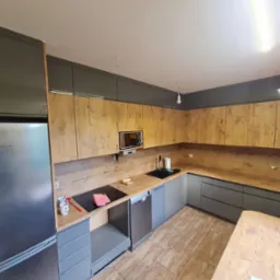 Nowoczesna kuchnia z szarymi szafkami dolnymi i drewnopodobnymi górnymi, srebrna lodówka Bosch po lewej, płyta indukcyjna z czerwoną ściereczką, jasna podłoga z płytek imitujących drewno.