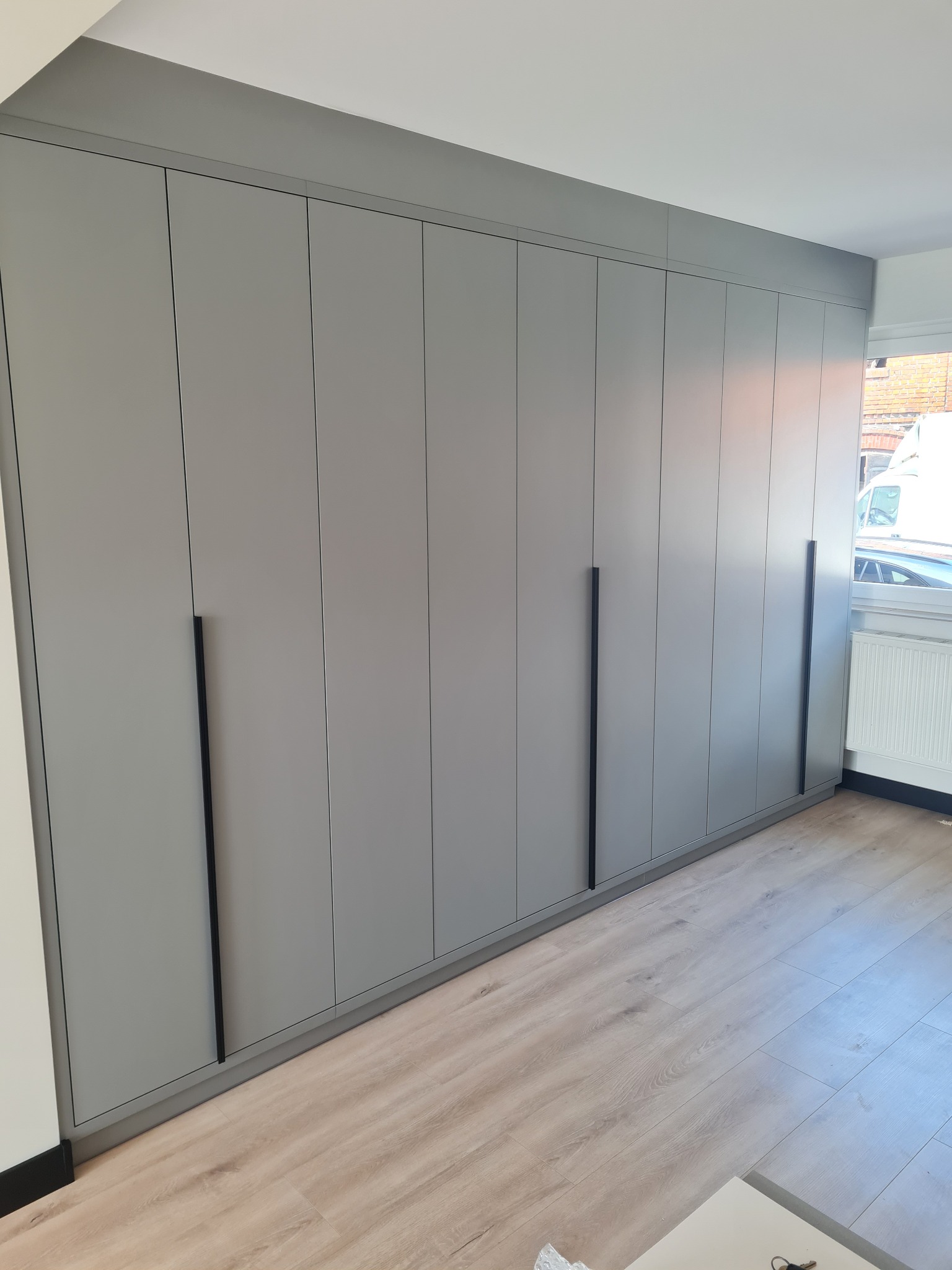 Szara zabudowa meblowa od podłogi do sufitu z minimalistycznymi, pionowymi uchwytami w czarnym kolorze, na tle jasnej podłogi imitującej drewno i okna z widokiem na ulicę.