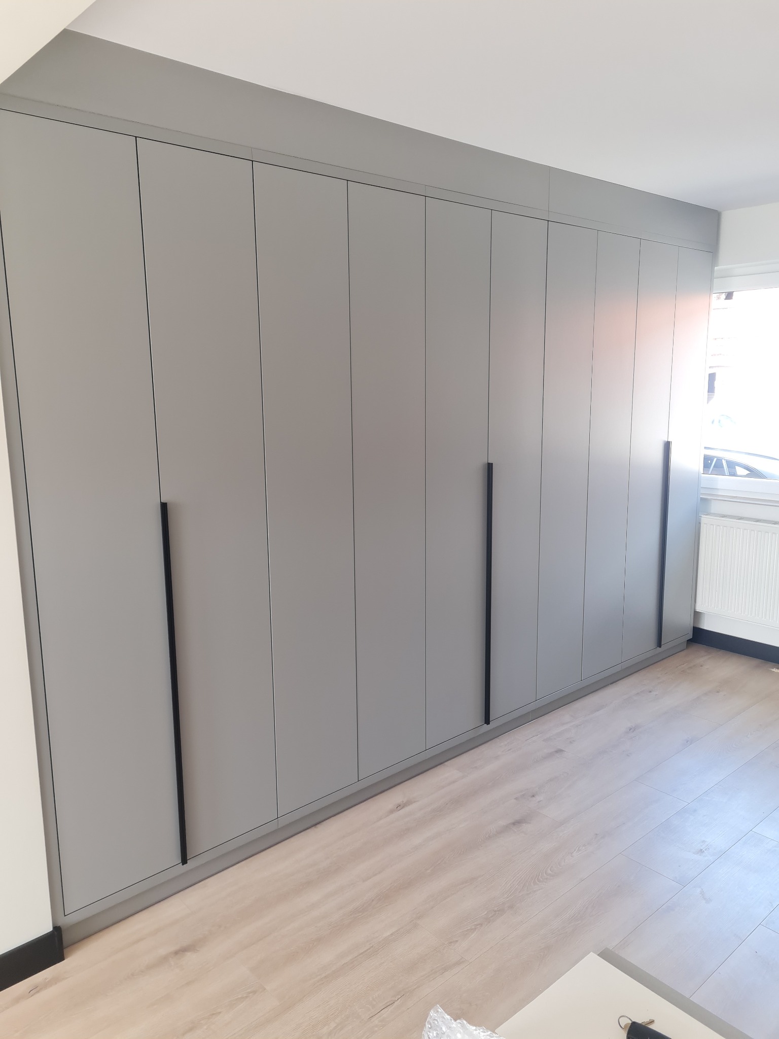 Szara szafa wnękowa z długimi, czarnymi uchwytami, dopasowana do sufitu, na podłodze panele imitujące jasne drewno, widok z dołu.