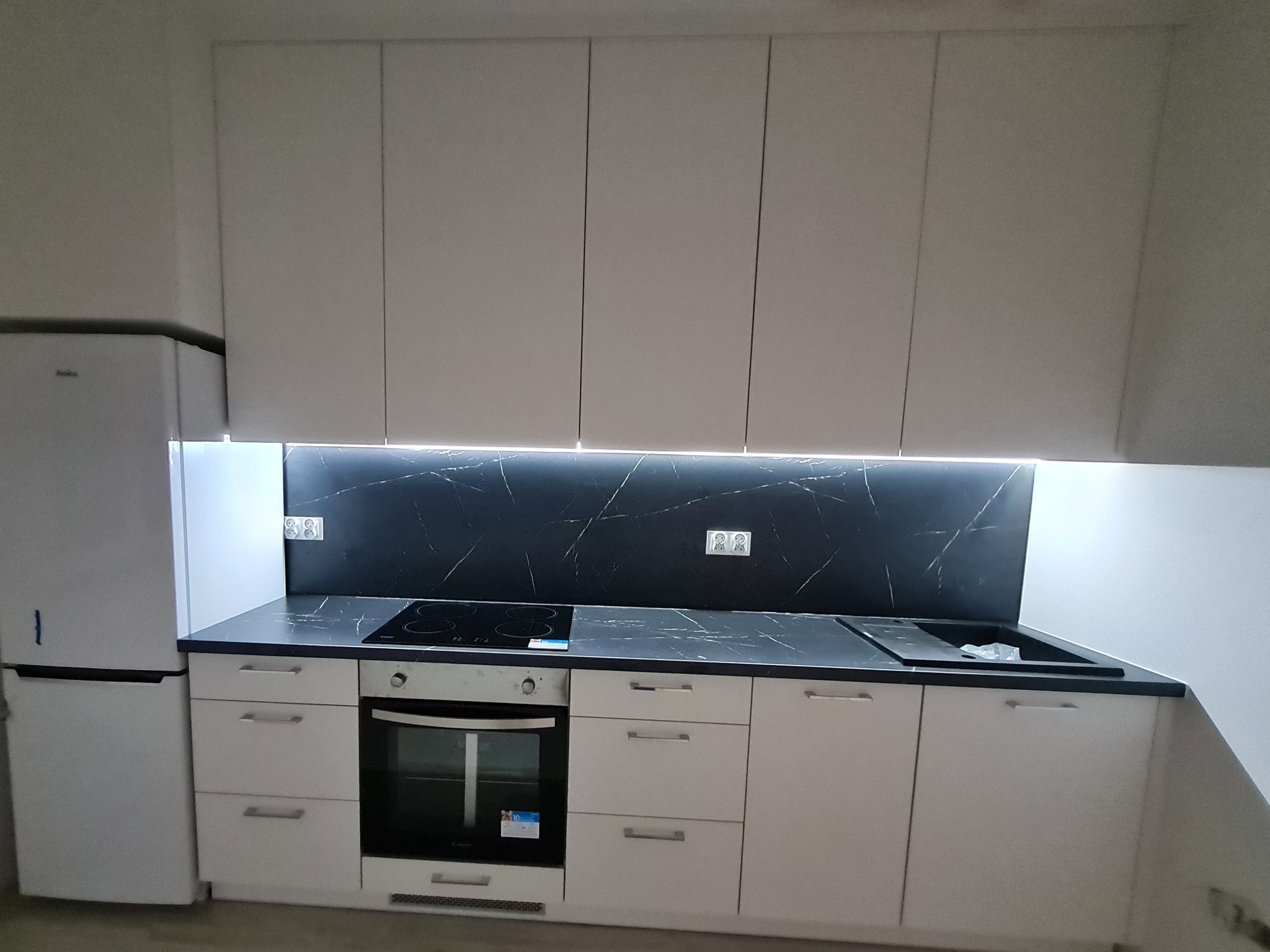 Nowoczesna, minimalistyczna kuchnia z białymi frontami szafek, czarnym blatem i panelem ściennym imitującym marmur, oświetlenie LED pod szafkami wiszącymi, widoczna płyta indukcyjna i zlewozmywak.