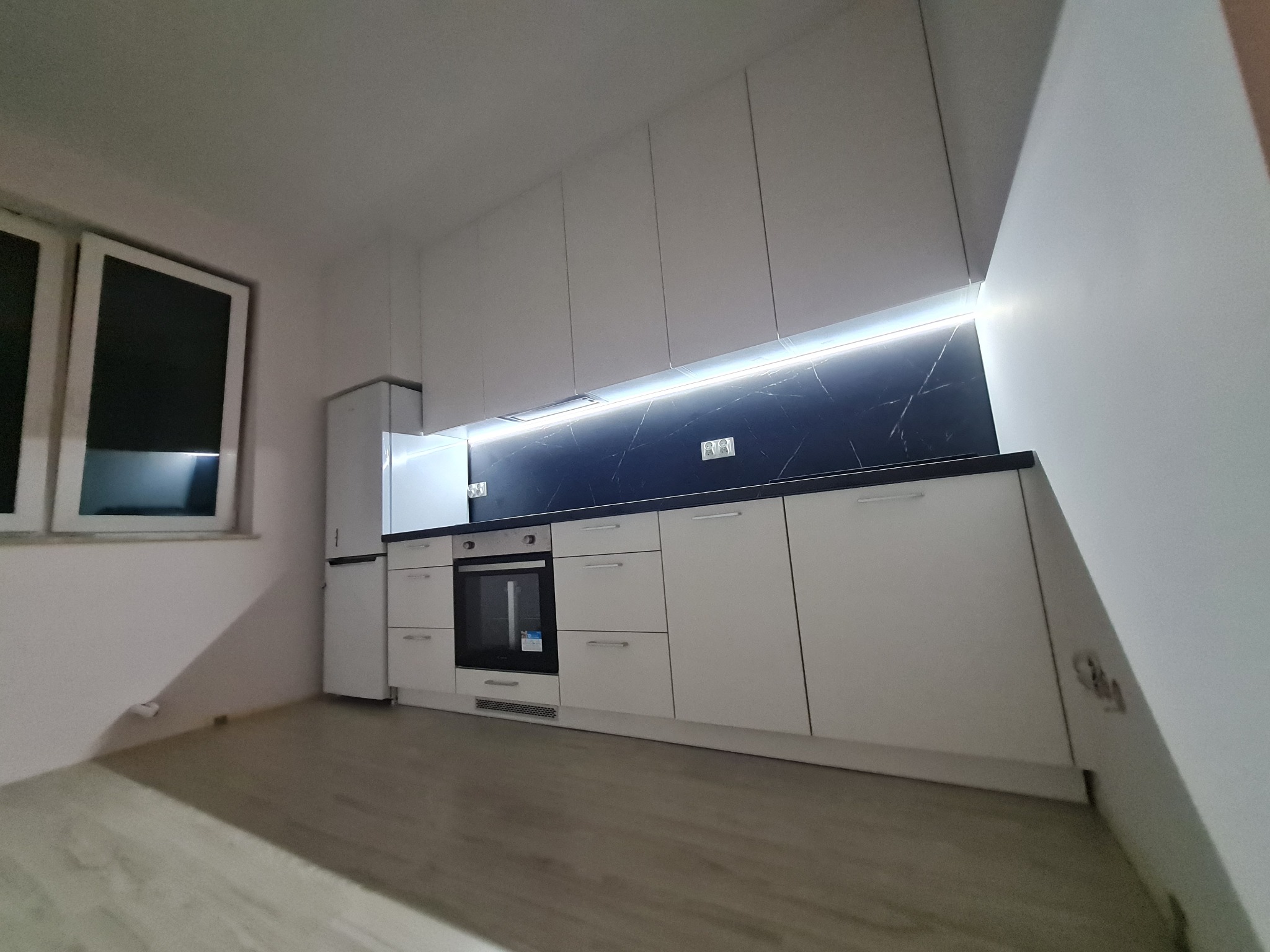 Nowoczesna, minimalistyczna kuchnia z białymi frontami, czarnym blatem i podświetleniem LED, widok z niskiej perspektywy na tle jasnego pomieszczenia z oknem.