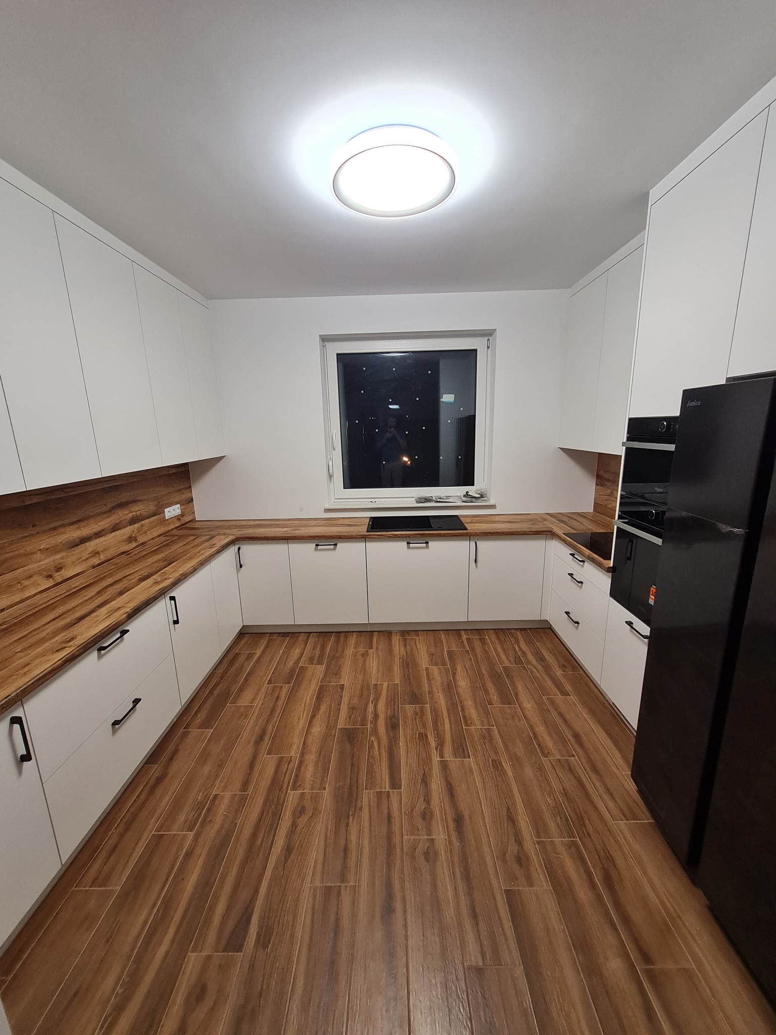 Nowoczesna kuchnia w kształcie litery U z białymi szafkami, drewnianym blatem i podłogą imitującą drewno, oświetlona centralnym plafonem.