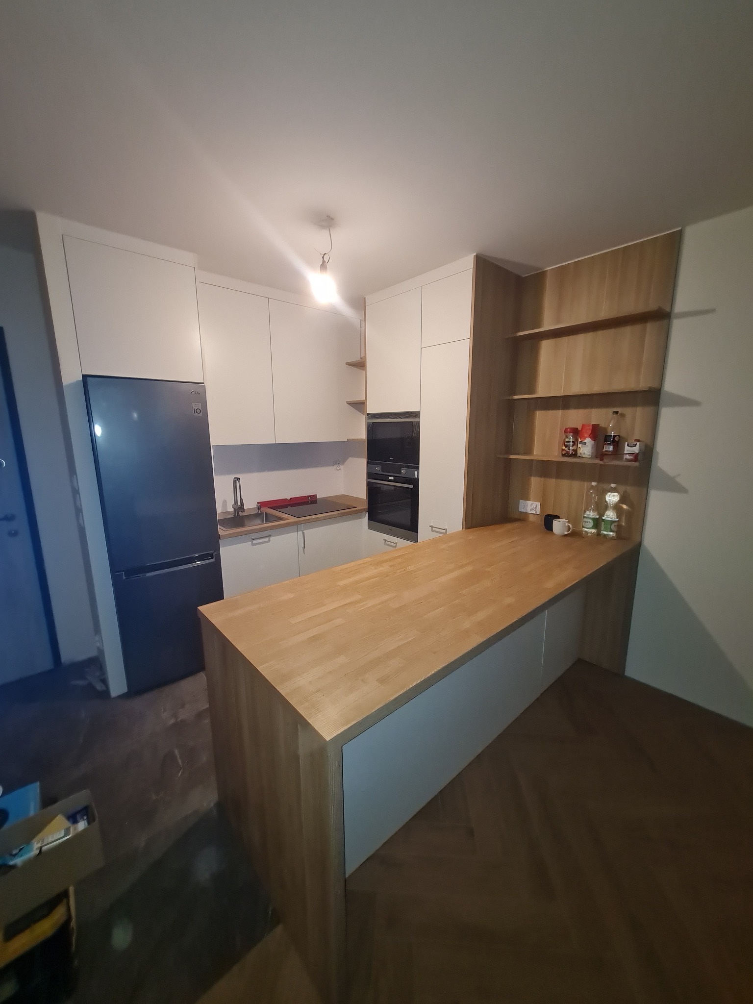 Nowoczesna kuchnia z białymi szafkami, drewnianym blatem i wyspą kuchenną, widok z perspektywy podłogi, z otwartymi półkami na ścianie. Na blacie stoją przyprawy i napoje.