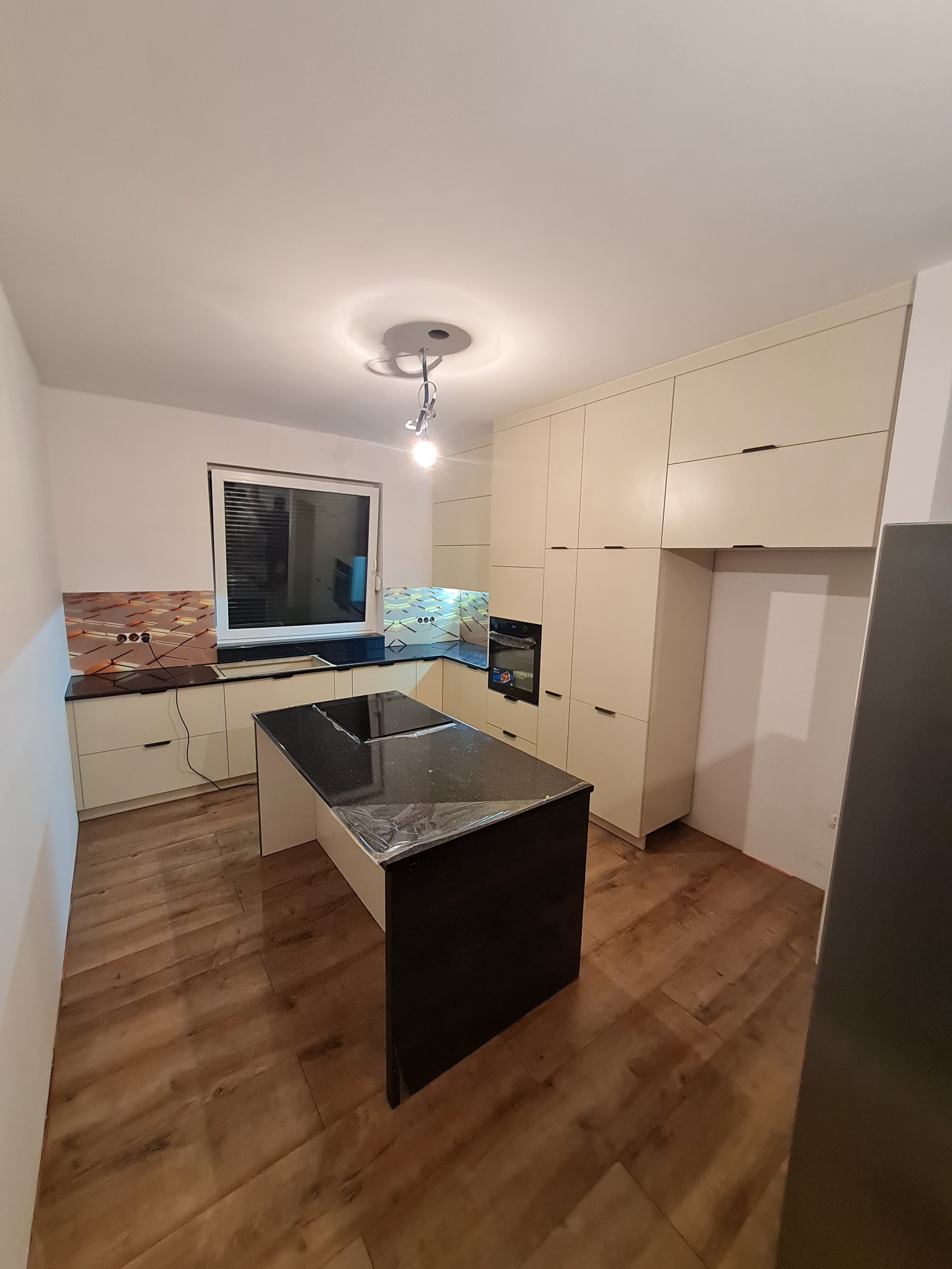 Nowoczesna kuchnia z wyspą i beżowymi szafkami, czarny blat wyspy zabezpieczony folią, parkiet na podłodze.