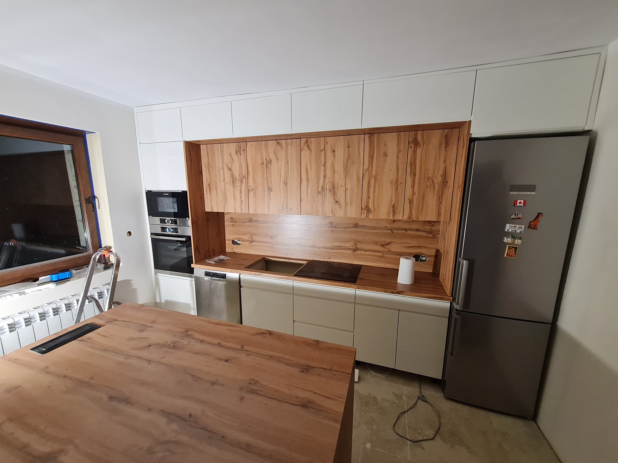 Wnętrze kuchni w trakcie montażu: białe szafki górne, drewnopodobne szafki wiszące i blat, sprzęt AGD w zabudowie, drabina przy oknie.