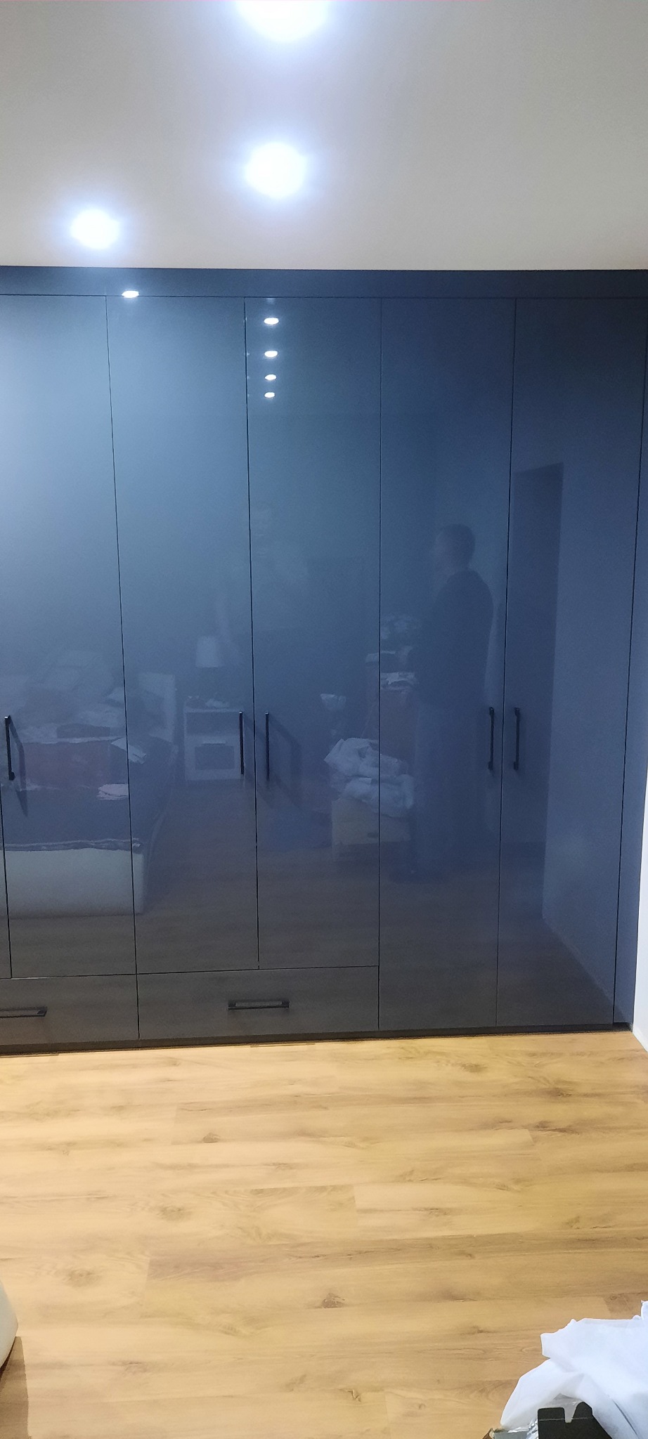 Szafa wnękowa z ciemnymi, lakierowanymi frontami, widoczne odbicie sylwetki osoby, szuflady w dolnej części, parkiet na podłodze.