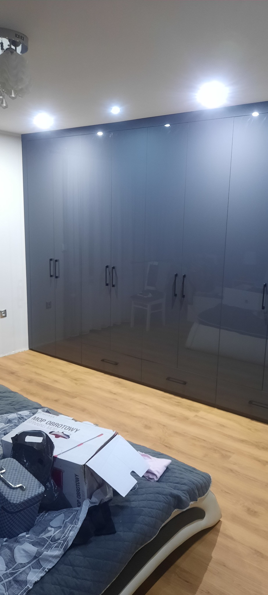 Duża, ciemnoszara szafa wnękowa z połyskującymi frontami i podłużnymi uchwytami, widoczna w sypialni z łóżkiem zagraconym pudełkami i przedmiotami.