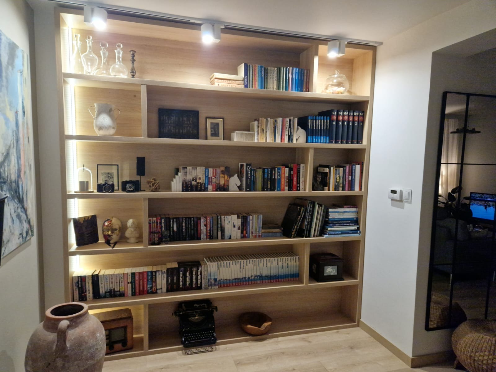 Jasna, drewniana biblioteczka na wymiar z oświetleniem punktowym, wypełniona książkami, dekoracjami i antykami, w tym maską wenecką i starą maszyną do pisania.