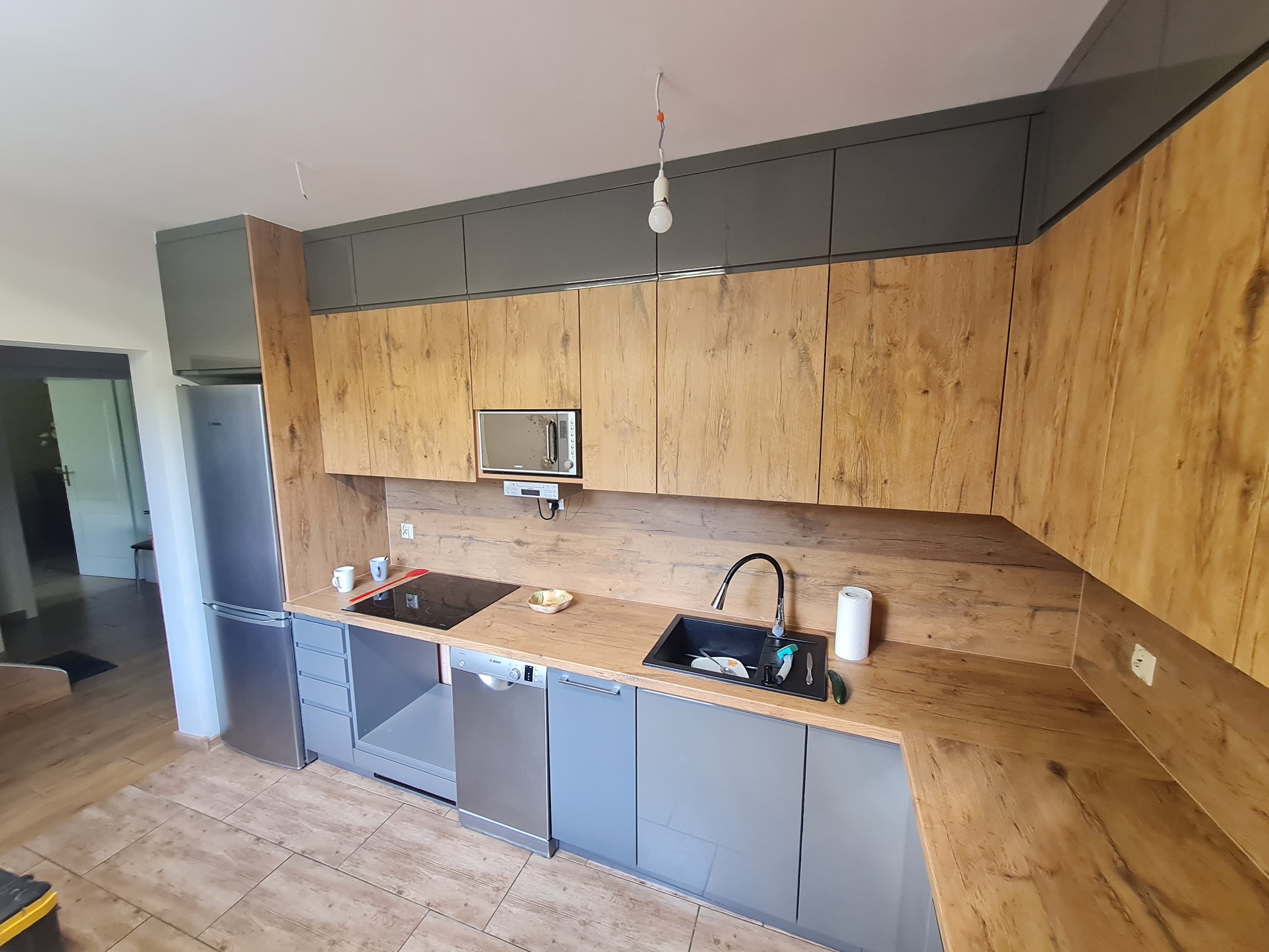 Nowoczesna kuchnia z drewnopodobnymi frontami szafek górnych i szarymi frontami dolnymi, widoczny zlew, płyta indukcyjna i sprzęt AGD, w tle niedokończone podłączenie oświetlenia.